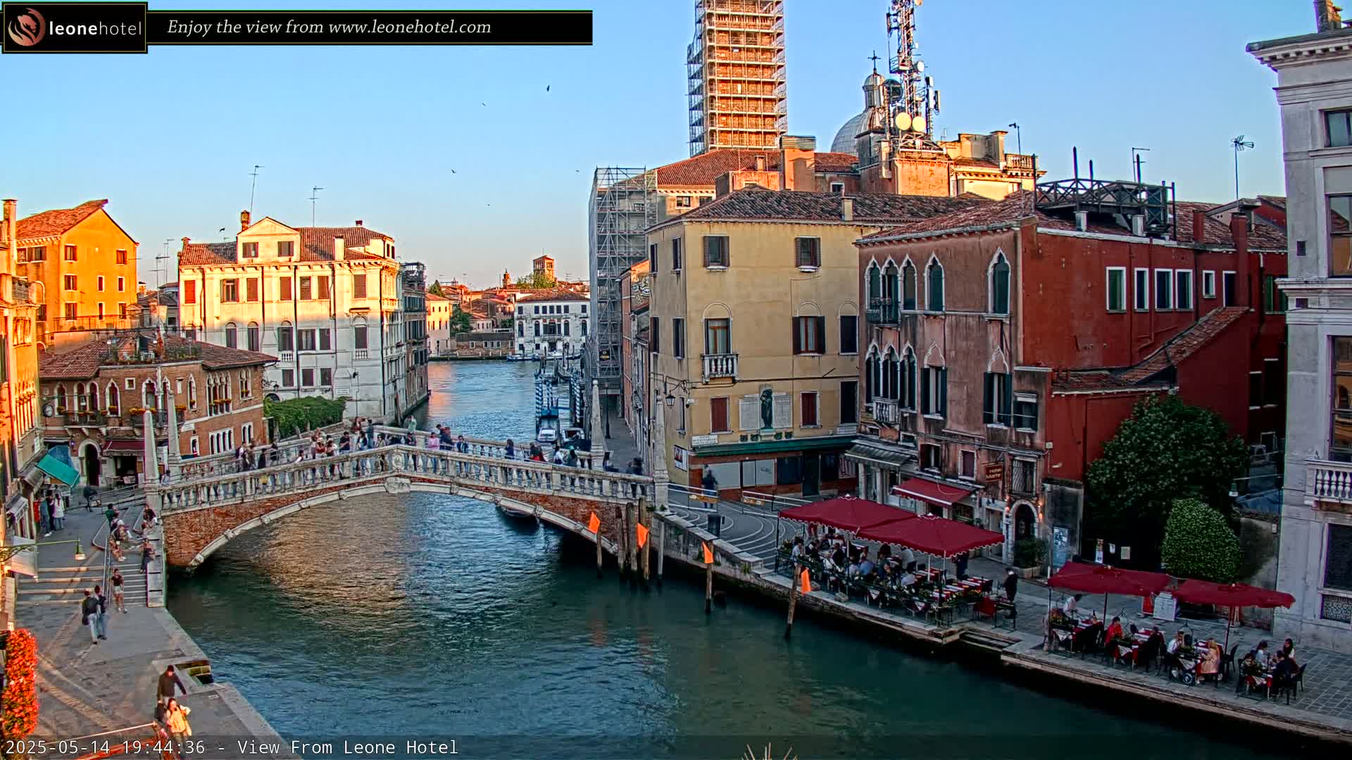 A canal in Venice, Italy, at sunset, shows several stone bridges, buildings, and outdoor seating areas along the waterway under a clear sky.