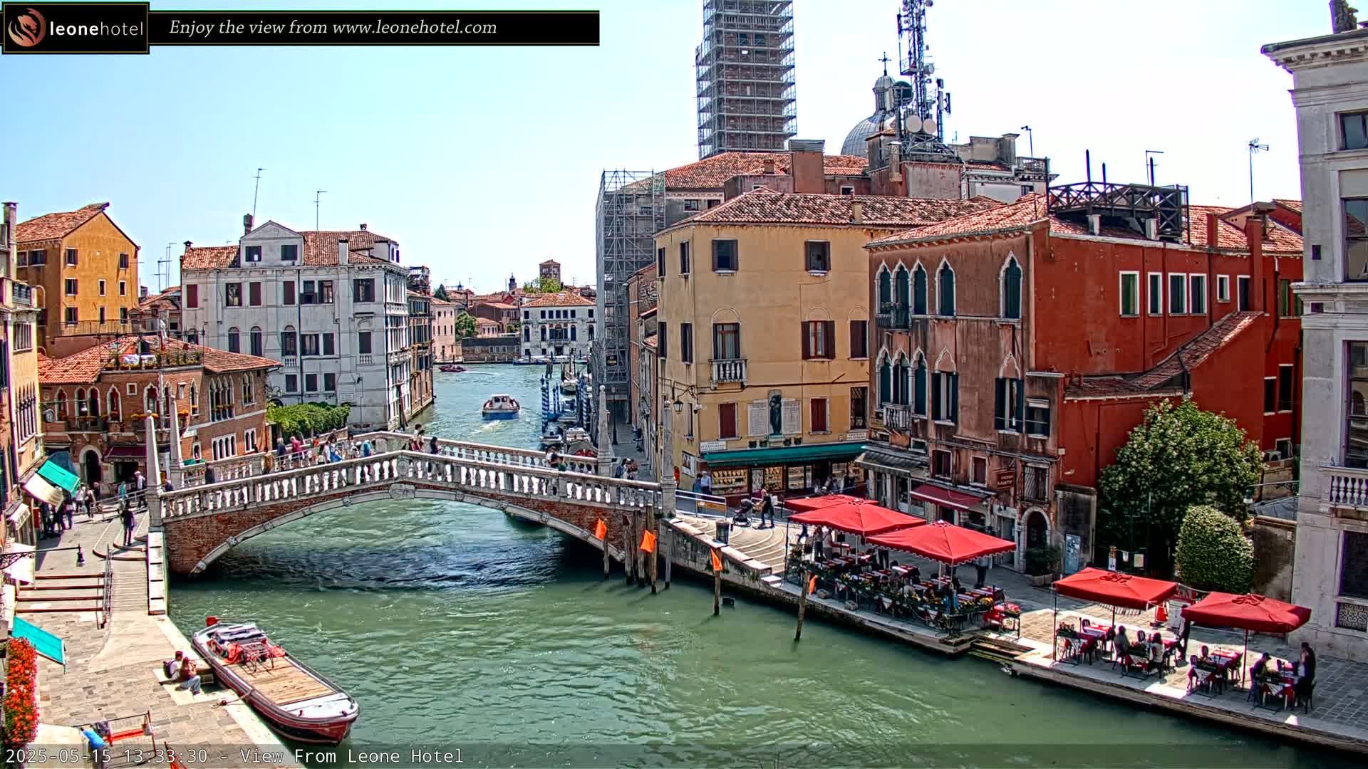 A sunny view of a canal in Venice, Italy, shows several buildings lining the waterway, a bridge crossing the canal, and outdoor seating areas at restaurants.