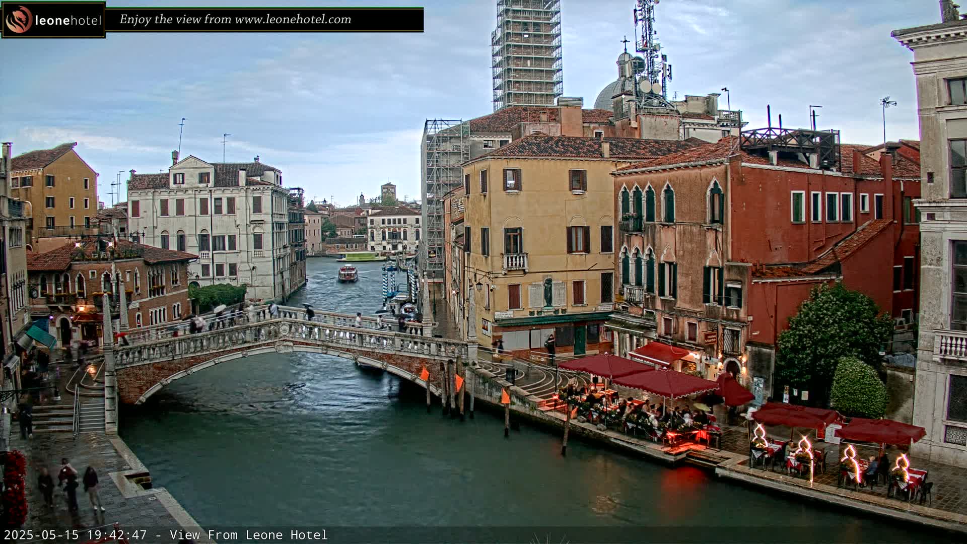 A stone bridge arches over a canal in Venice, Italy, under a cloudy sky;  several buildings line the canal, some with outdoor seating areas, and a boat is visible on the water.
