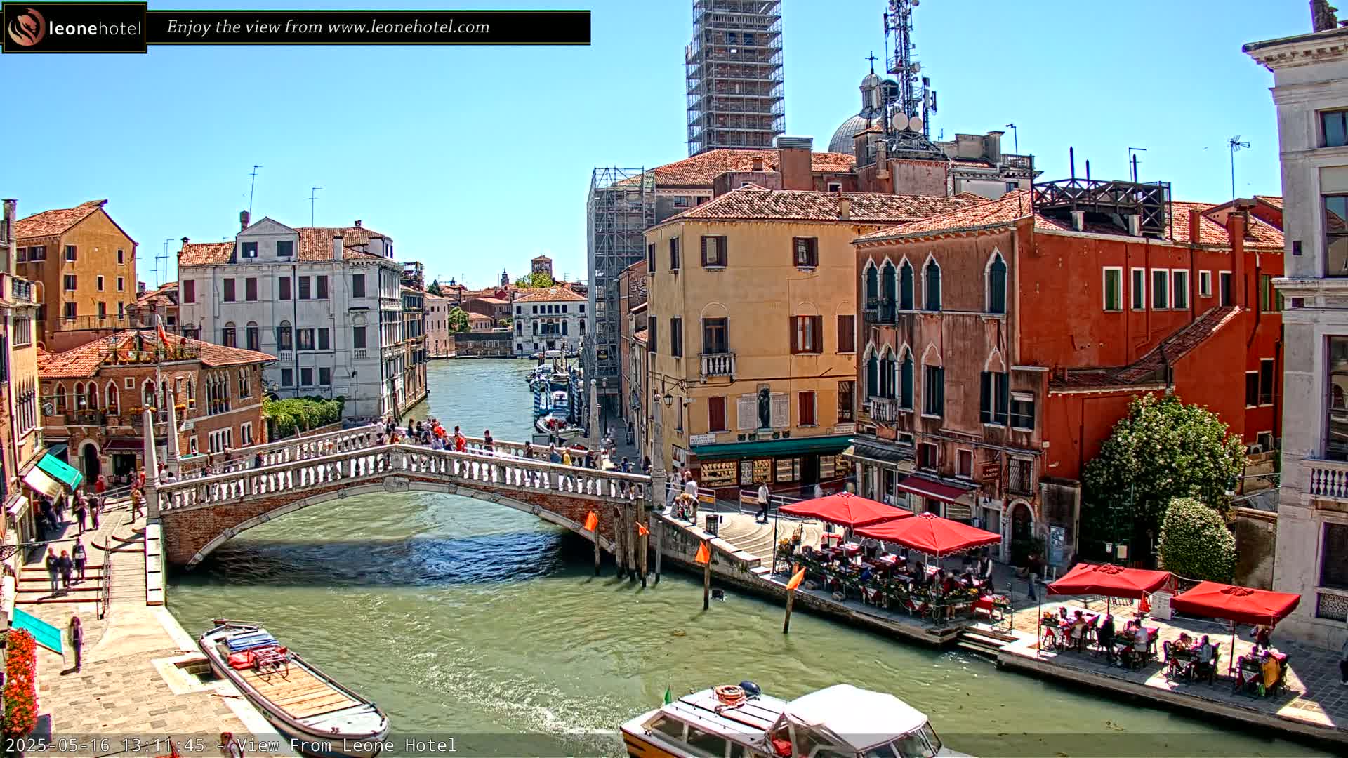 A sunny day in Venice shows a canal with a stone bridge, colorful buildings lining the waterway, and outdoor seating at cafes.