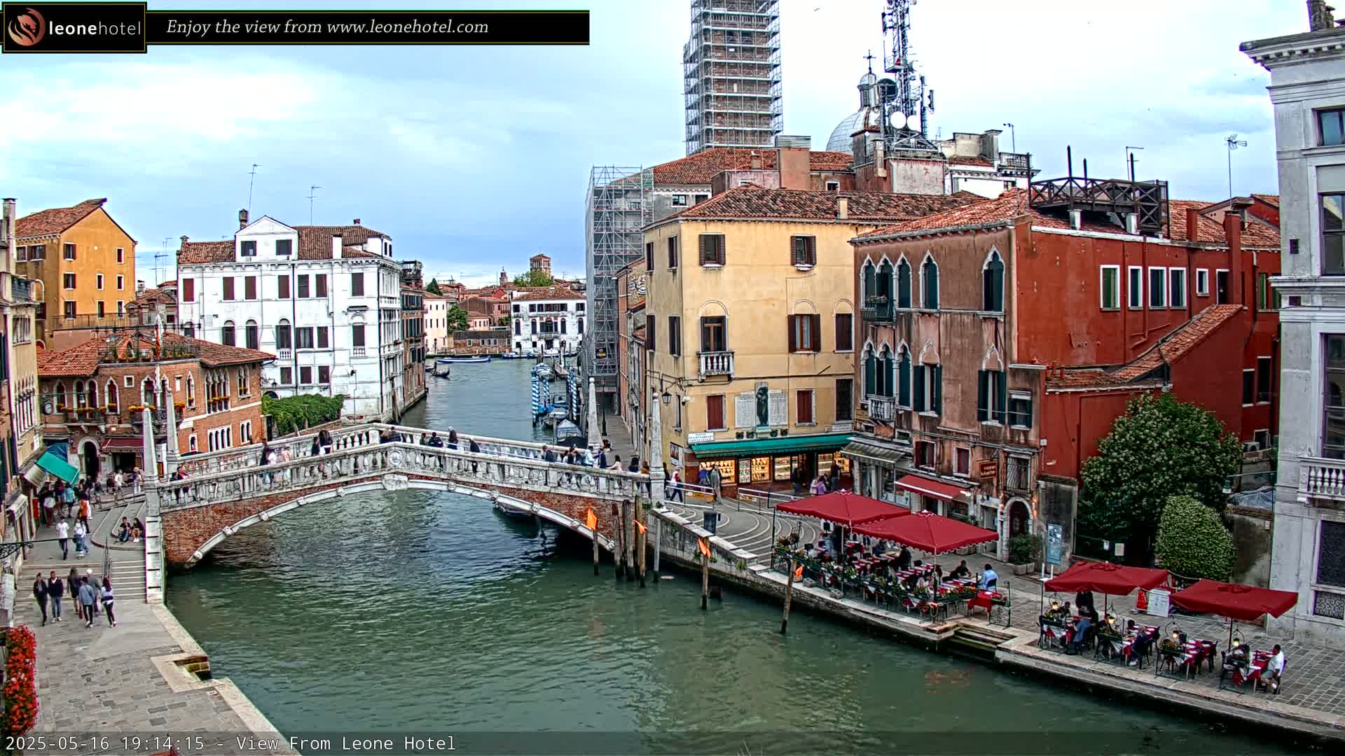 A stone bridge spans a canal in Venice, Italy, under a partly cloudy sky; colorful buildings line the waterway, and people are walking on the bridge and dining outdoors at canalside restaurants.