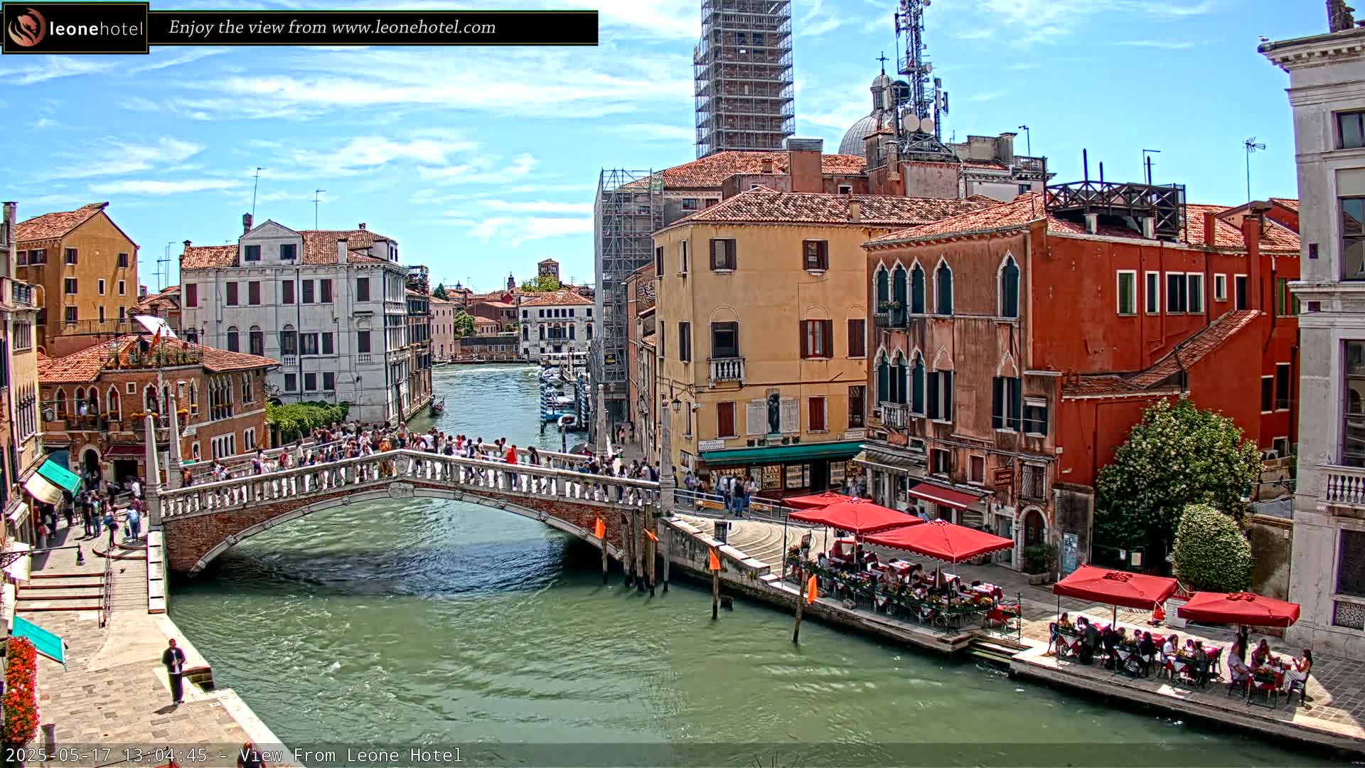 A sunny day in Venice shows a canal with a bridge crowded with people, colorful buildings lining the waterway, and outdoor seating at cafes along the canal.
