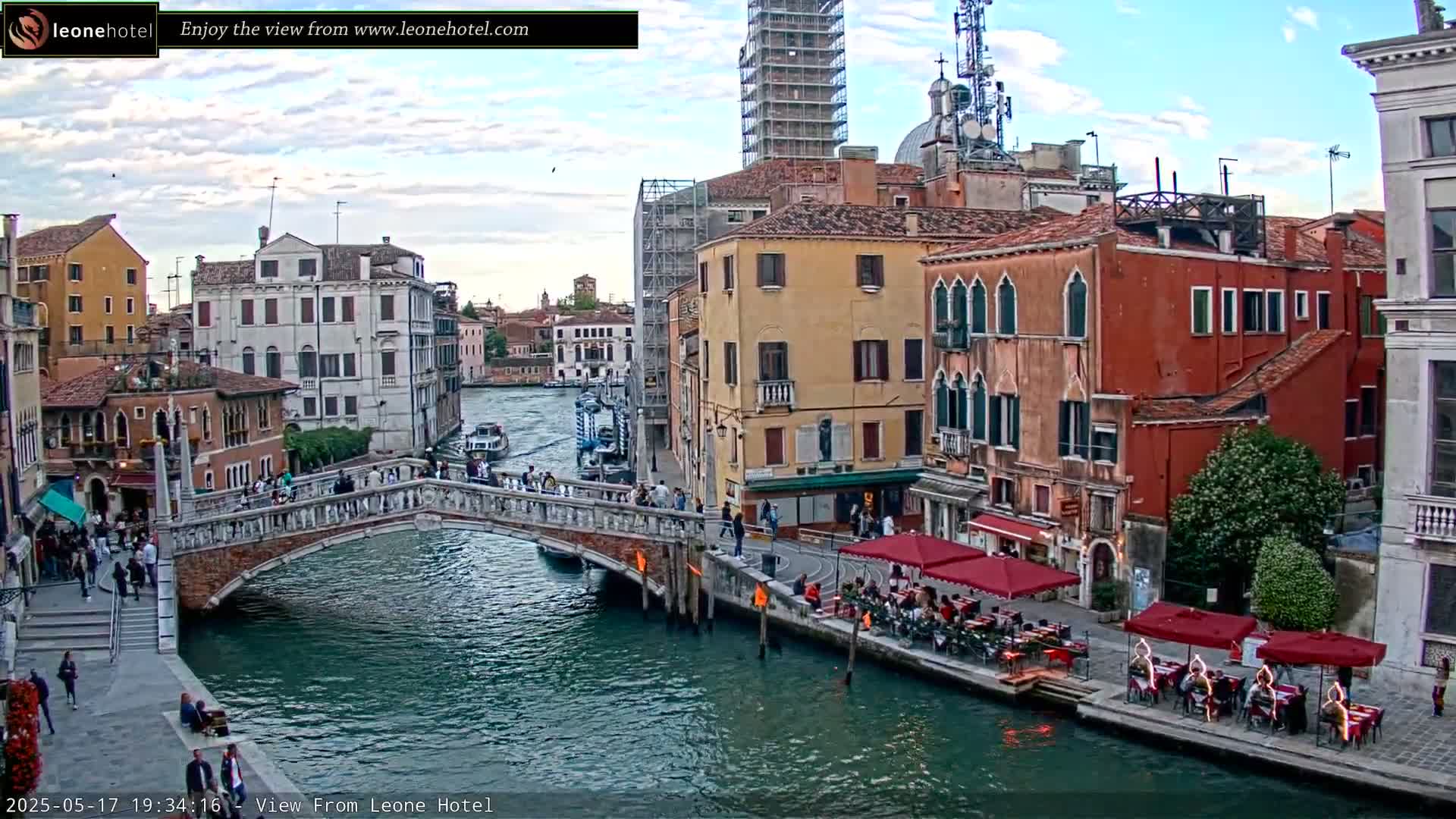 A Venetian canal scene under a partly cloudy sky shows a bridge with pedestrians, colorful buildings lining the waterway, and outdoor dining areas with patrons seated at tables under red umbrellas.