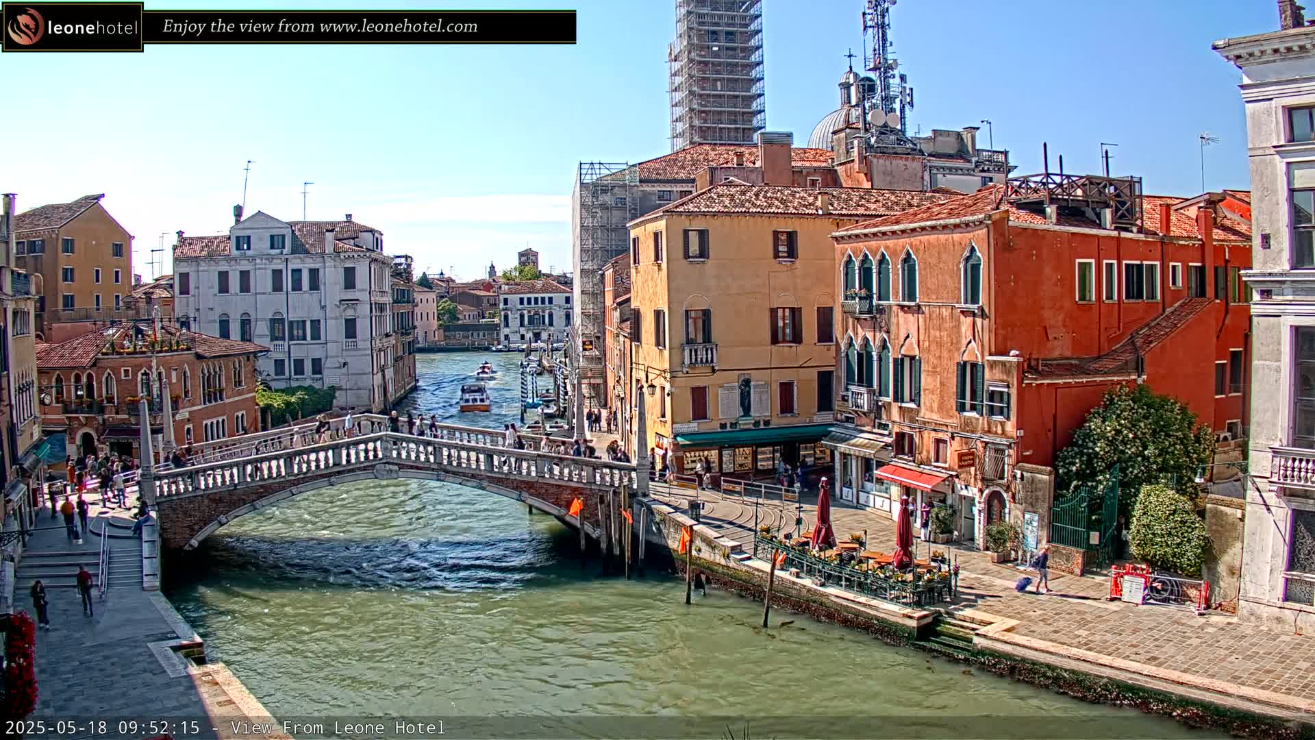 A sunny day in Venice shows a canal with a stone bridge, boats, and colorful buildings lining the waterway.