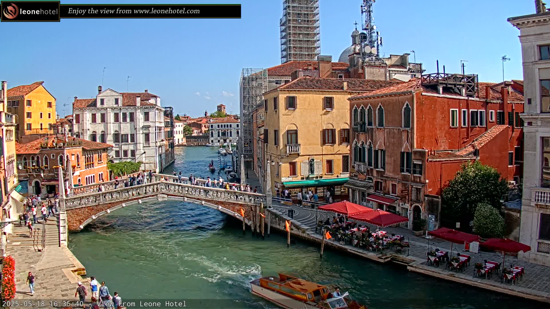 A sunny day in Venice shows a canal with a stone bridge crowded with people, colorful buildings lining the waterway, and several gondolas and motorboats.