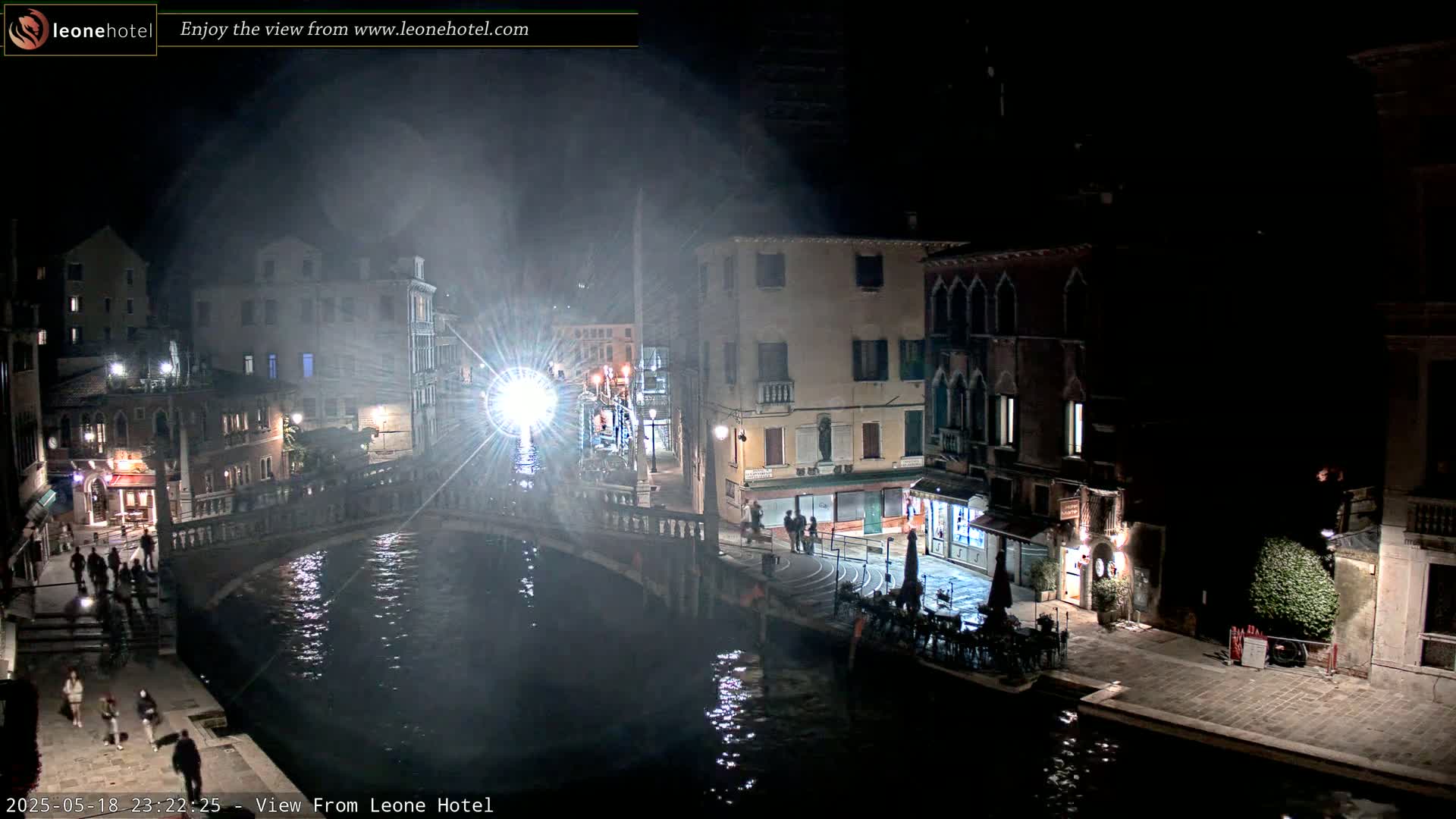 A nighttime view of a canal in Venice, Italy, showing several buildings along the waterway with some people walking around, under hazy or foggy conditions.