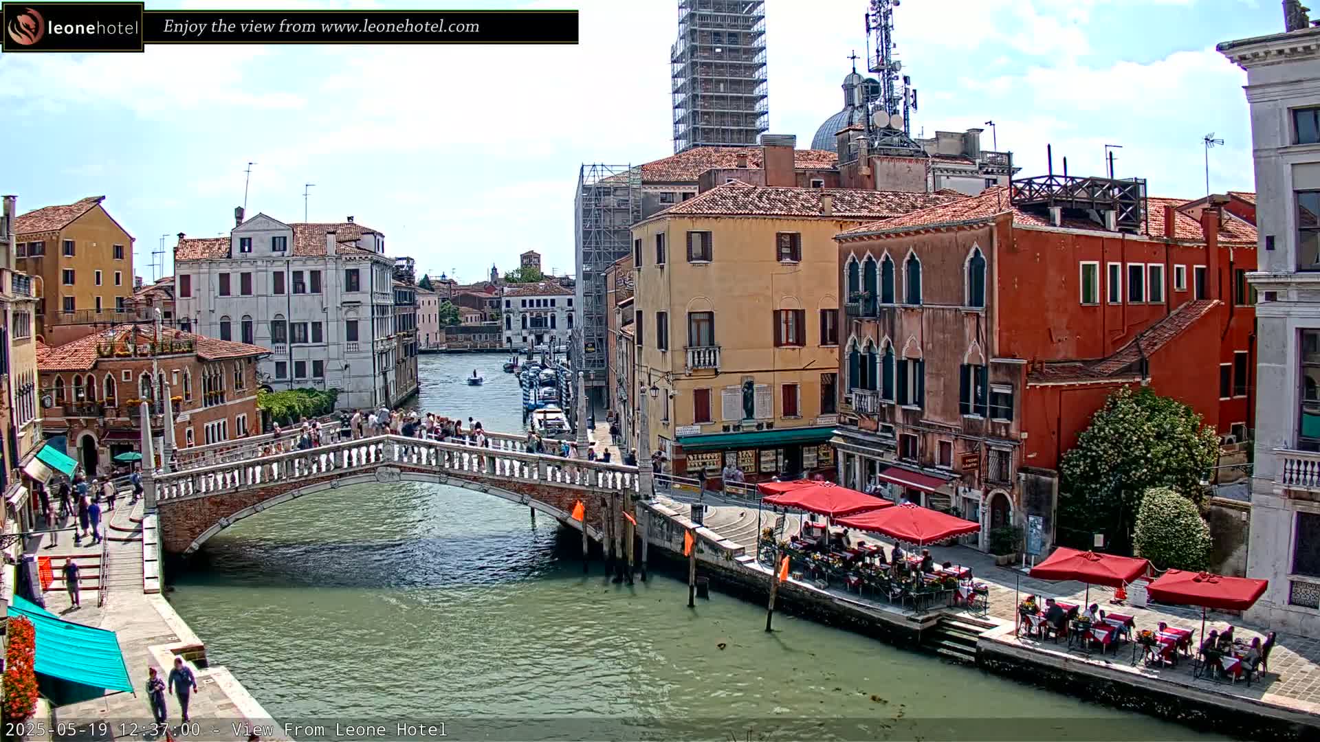 A sunny day in Venice shows a canal with a stone bridge and colorful buildings lining the waterway, with outdoor seating at cafés along the canal.