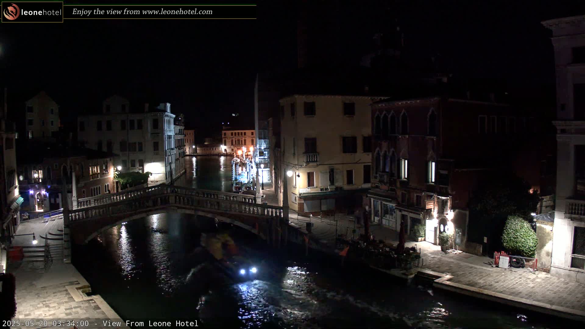 A nighttime view of a canal in Venice, Italy, shows a bridge, buildings lining the waterway, and a small motorboat with lights on the water.