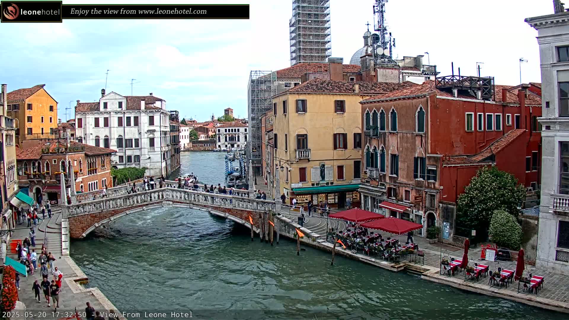 A canal in Venice, Italy, with several bridges and buildings along its banks, under partly cloudy skies, shows pedestrians walking across a stone bridge and patrons sitting at outdoor cafes.