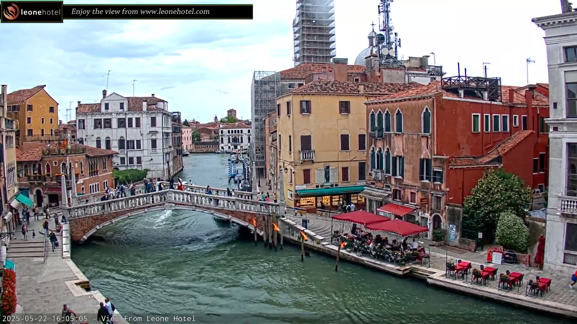 A canal in Venice, Italy, under a partly cloudy sky, is shown with a bridge, several buildings, and outdoor seating at a restaurant.