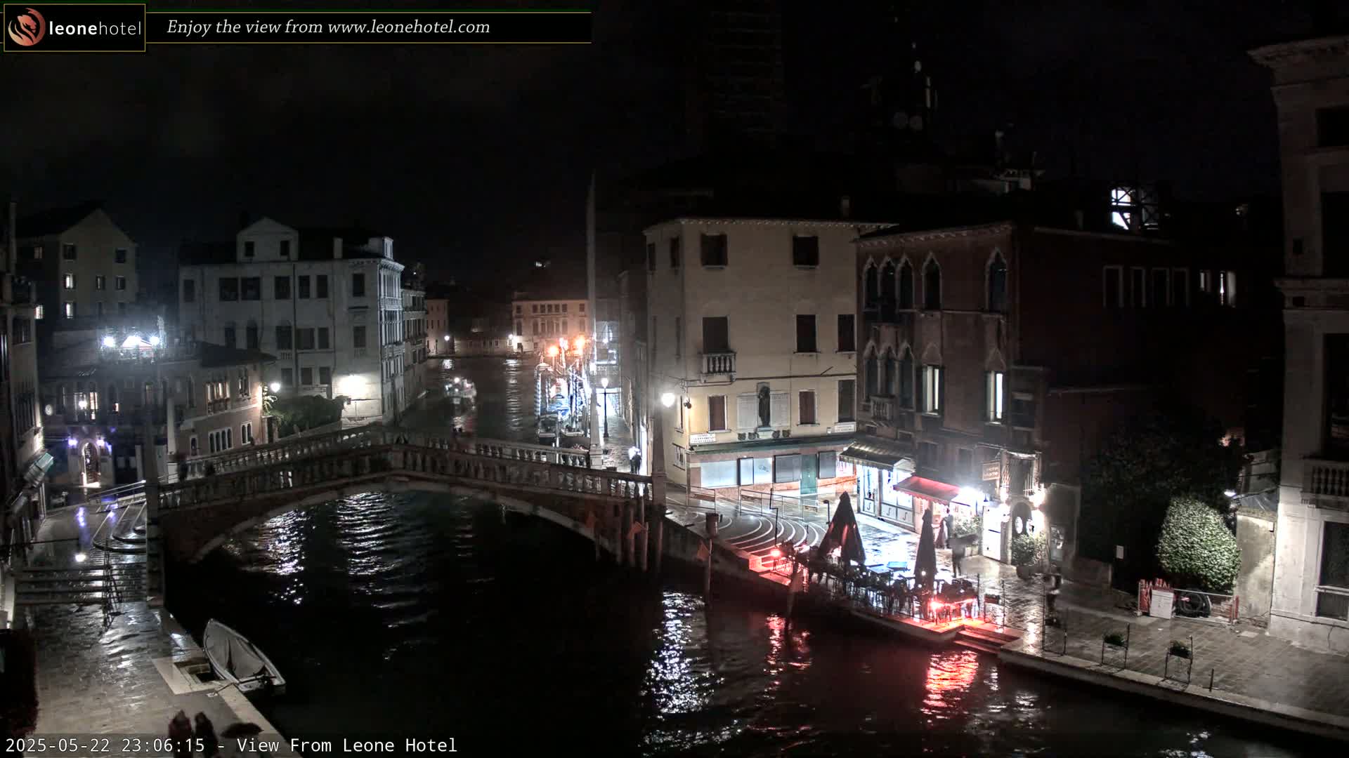A nighttime view of a canal in Venice, Italy, showing a bridge, buildings, and a boat, with rain on the ground.