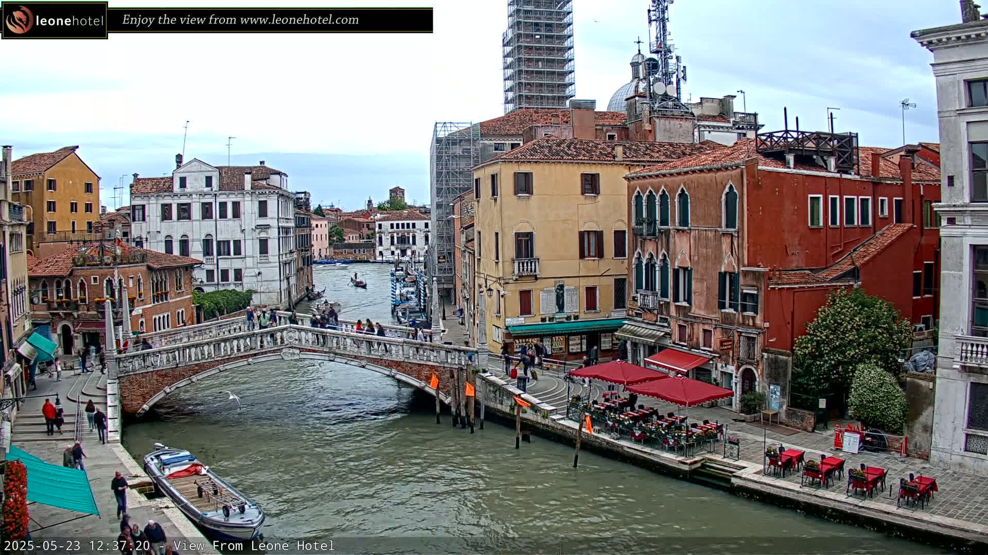 A canal in Venice, Italy, with overcast skies, shows several bridges, buildings, gondolas, and outdoor cafes along its banks.