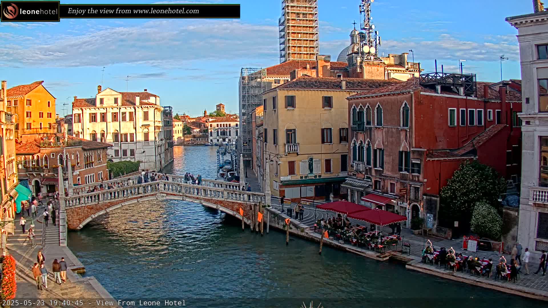 A stone bridge crosses a canal in Venice, Italy, under a mostly clear evening sky, with colorful buildings lining the waterway and people walking on the bridge and sitting at outdoor cafes.