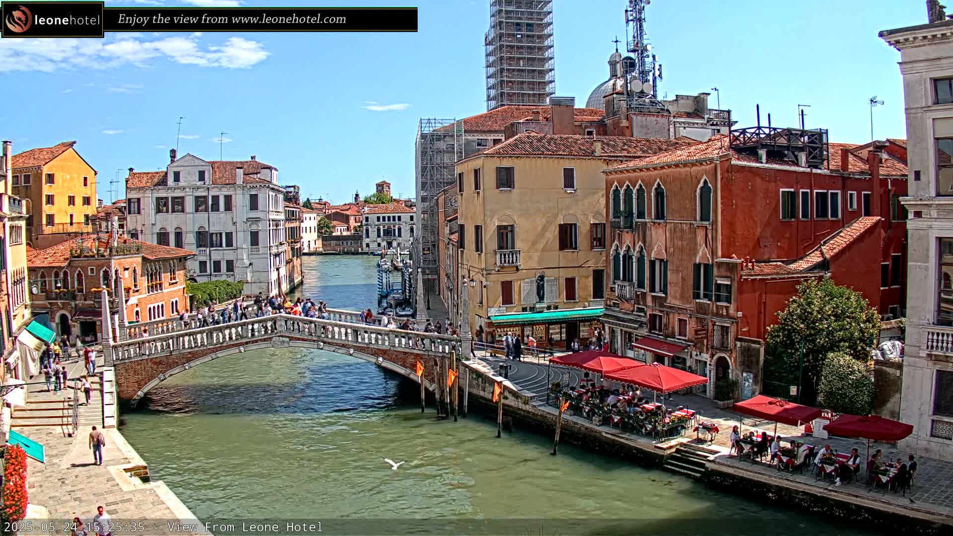 A sunny day in Venice shows a canal with a bridge, pedestrians, and waterfront restaurants under red umbrellas, all surrounded by historic buildings.