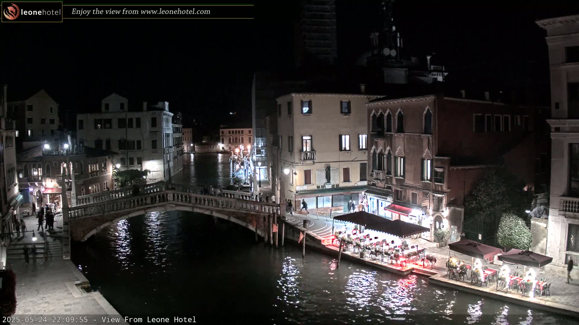 A nighttime view of a canal in Venice, Italy, shows a stone bridge, illuminated buildings, and outdoor dining areas along the water's edge.