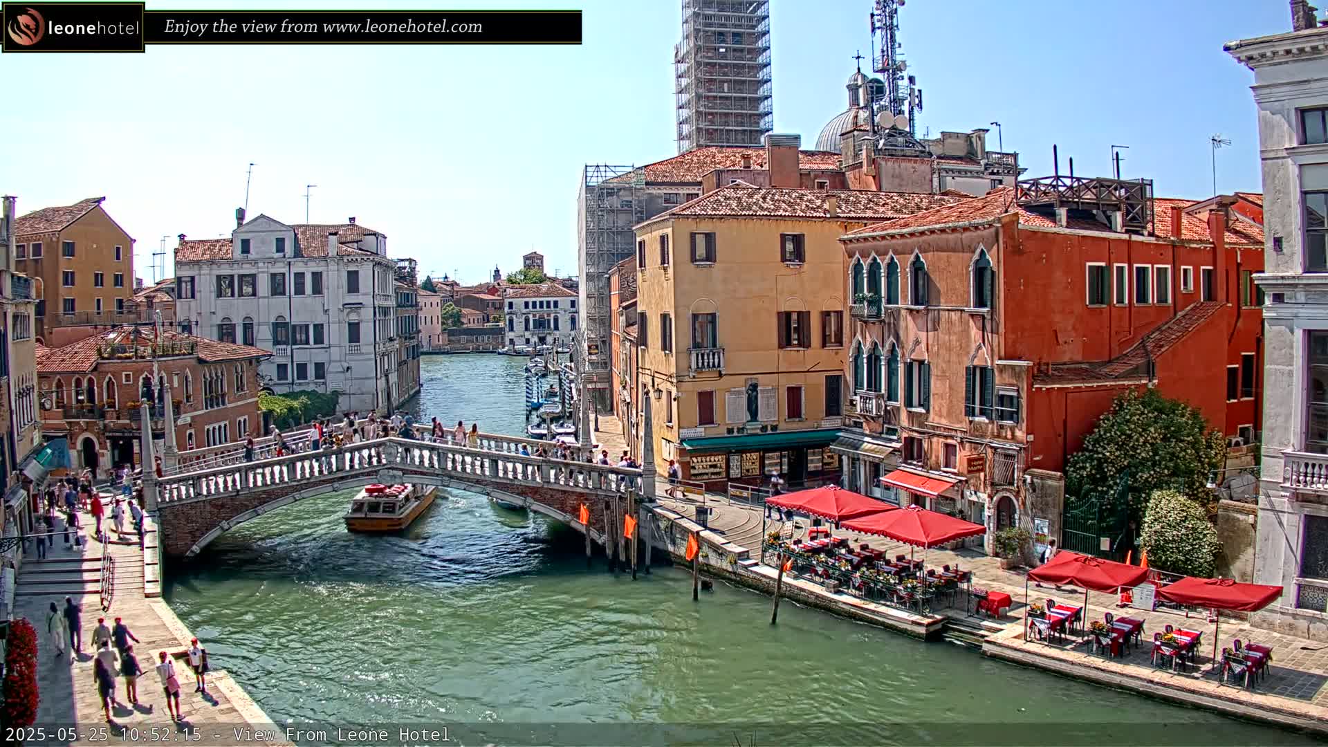 A sunny day in Venice shows a canal with a bridge, boats, and colorful buildings, with outdoor seating at cafes along the waterway.