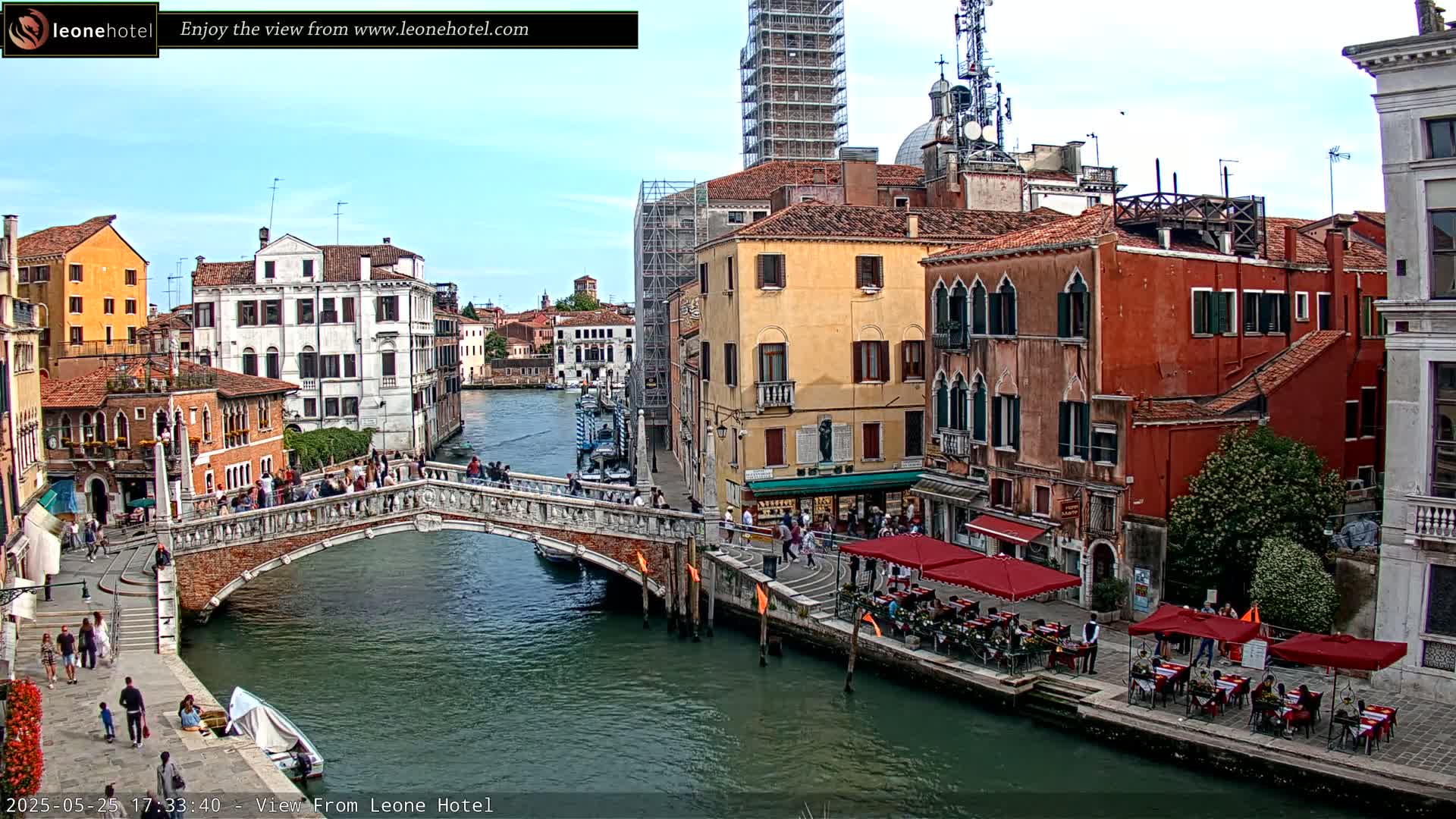 A sunny day in Venice shows a canal with a stone bridge, several buildings, and outdoor seating at a restaurant.