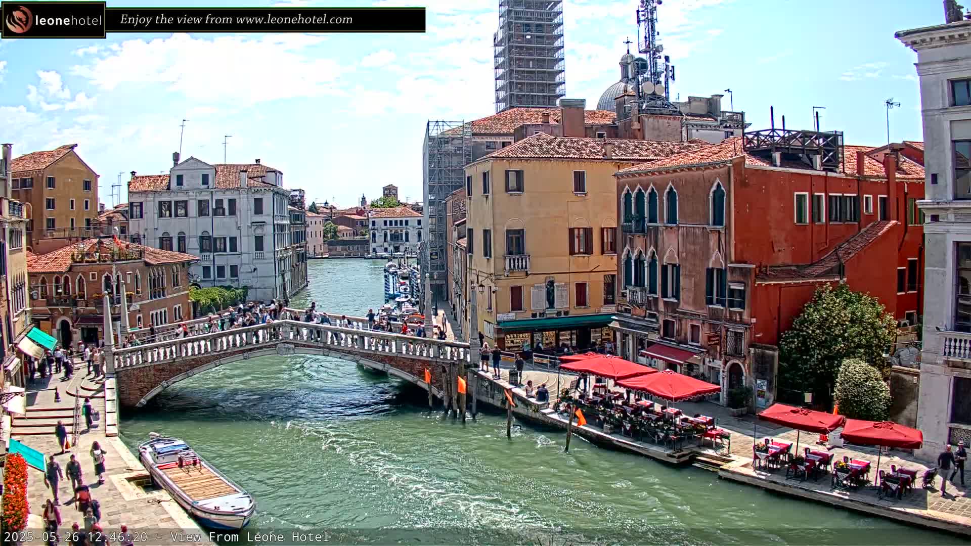 A sunny day in Venice shows a canal with a bridge crowded with people, boats moored along the sides, and outdoor cafes lining the waterfront.