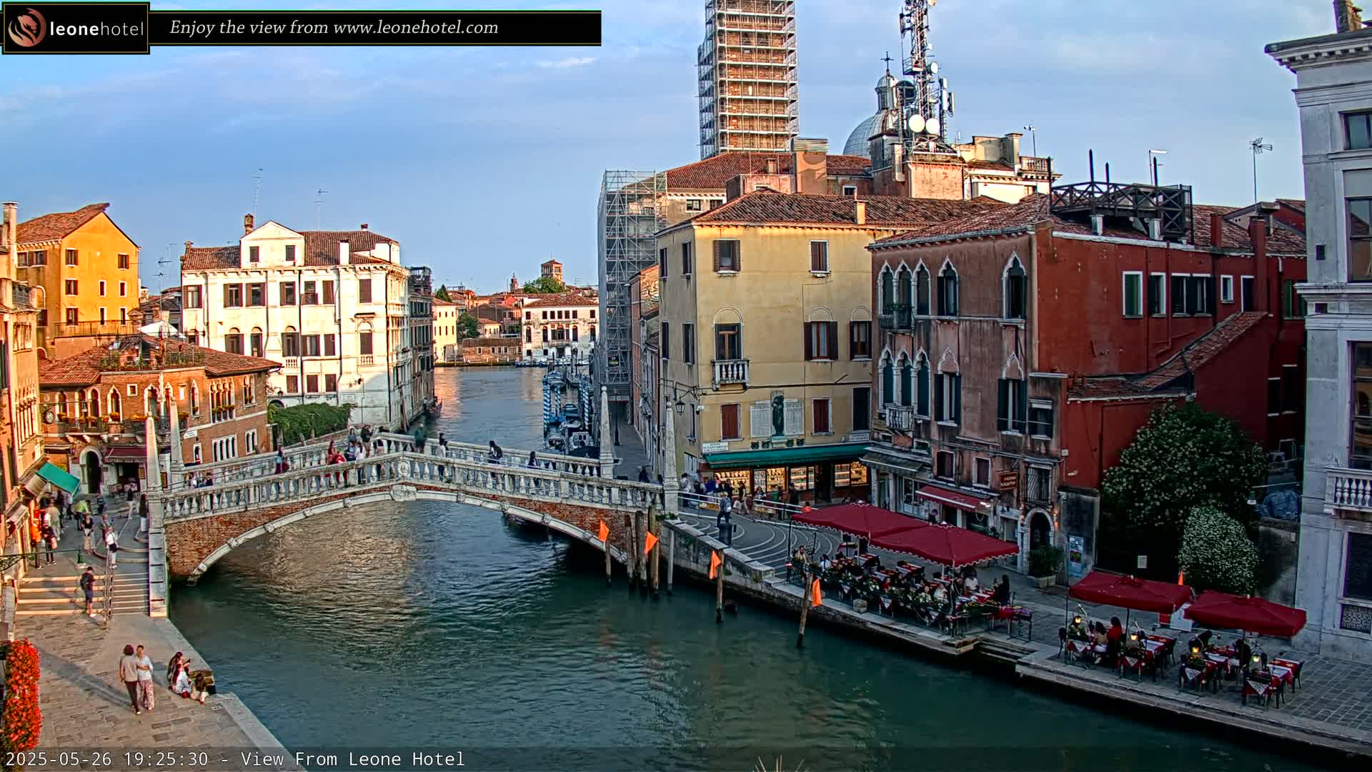 A stone bridge crosses a canal in Venice, Italy, under a mostly clear evening sky, with colorful buildings lining the waterway and outdoor seating at cafes along the canal.