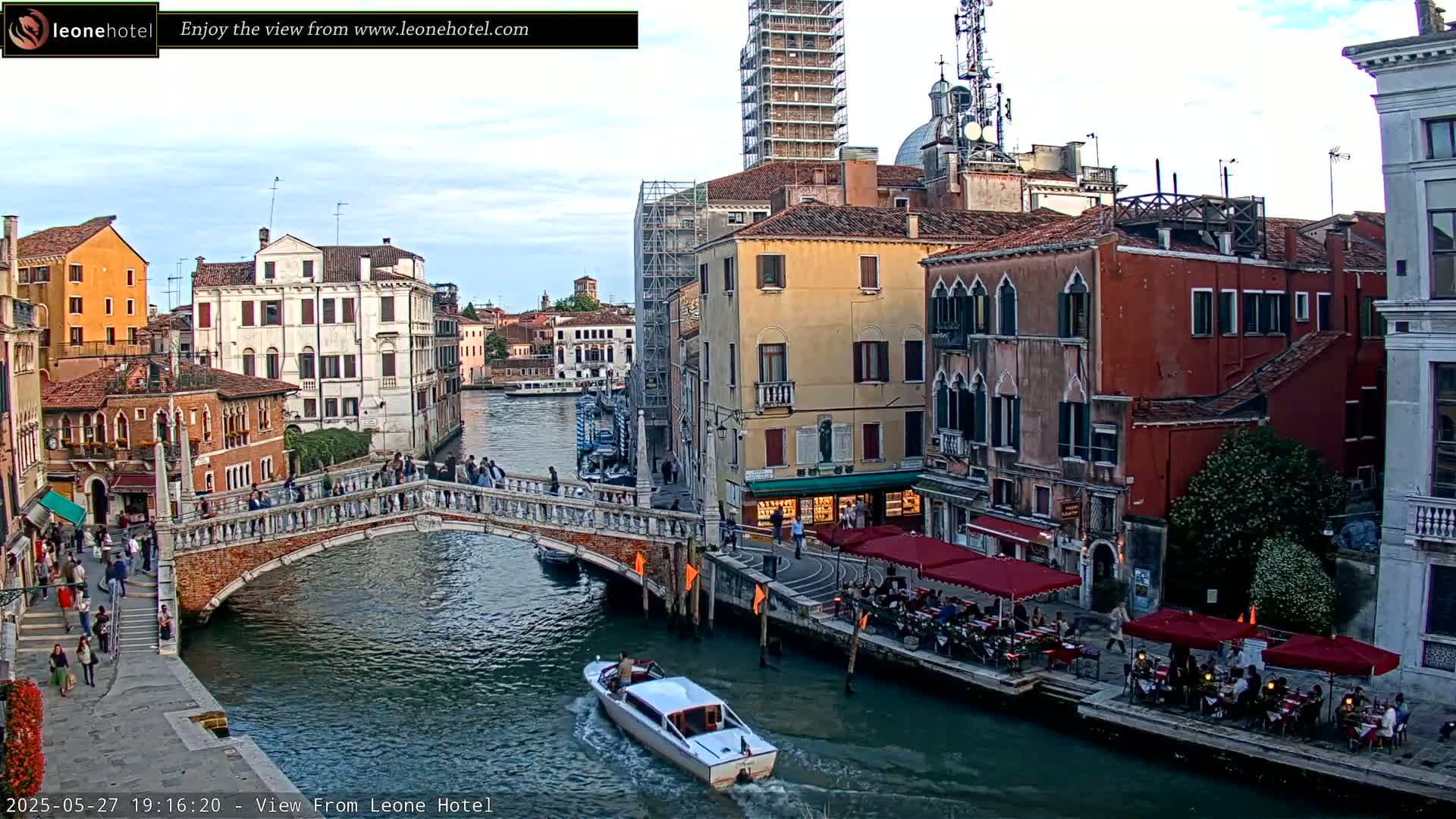 A canal in Venice, Italy, under a partly cloudy sky, is spanned by a stone bridge with people walking on it and along the canal's edge, where outdoor seating for restaurants lines the banks, and a motorboat travels on the water.