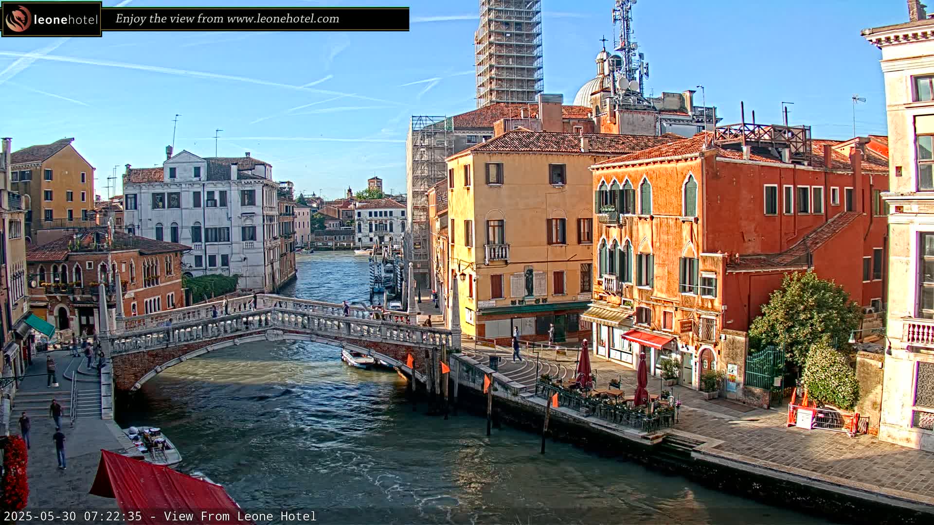 A canal in Venice, Italy, under a clear blue sky, is spanned by a stone bridge with people walking on it and colorful buildings lining its banks.