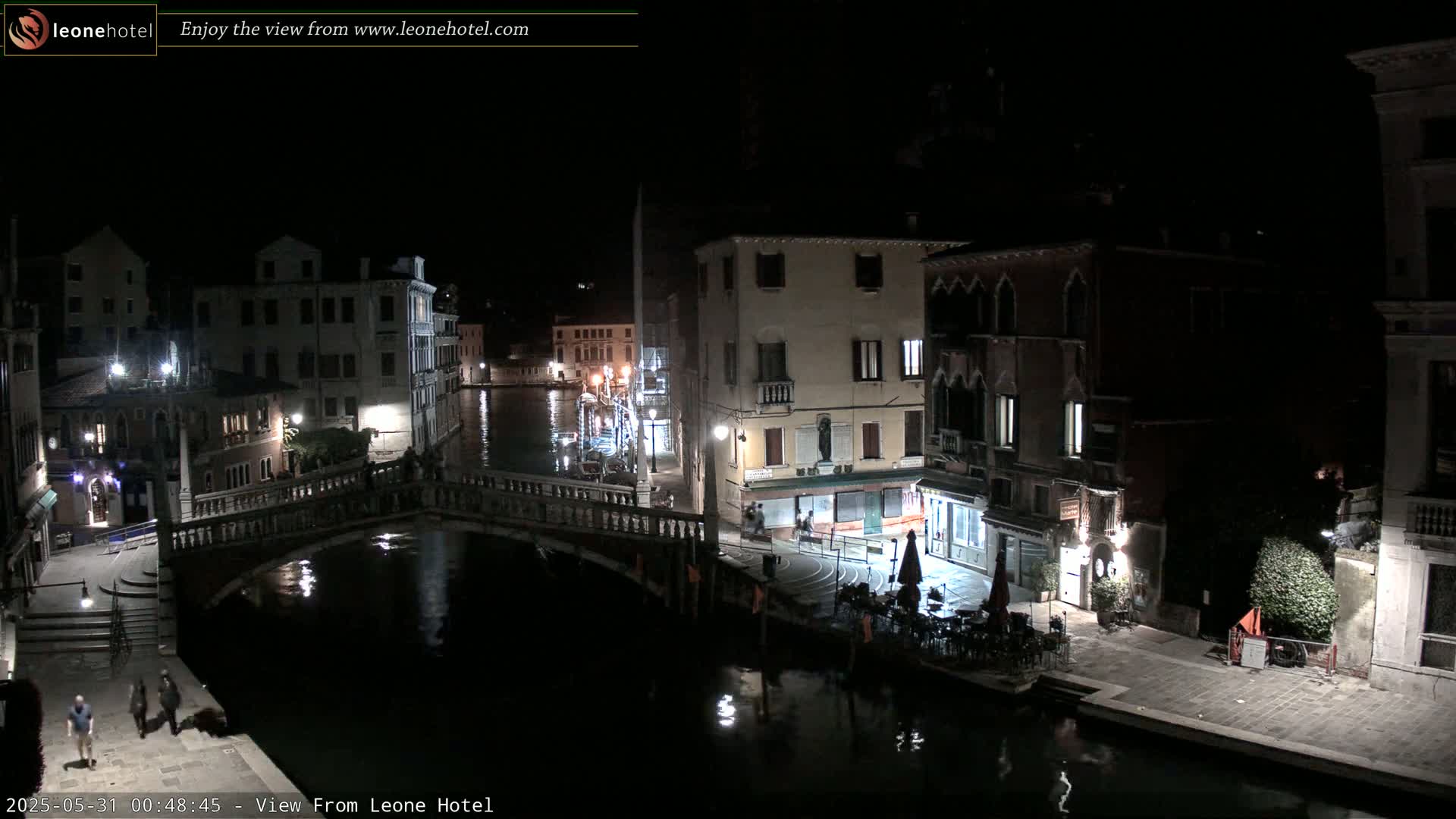 A nighttime view of a canal in Venice, Italy, shows several buildings lining the waterway, with a bridge crossing the canal and a few people walking along the waterfront.