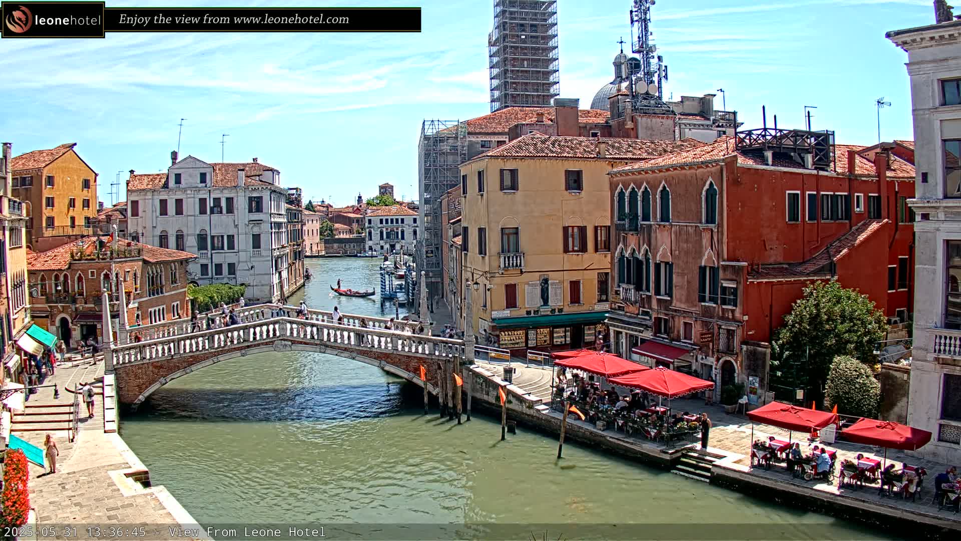 A sunny day in Venice shows a canal with a stone bridge, colorful buildings lining the waterway, and several outdoor cafes with patrons seated at red umbrellas.
