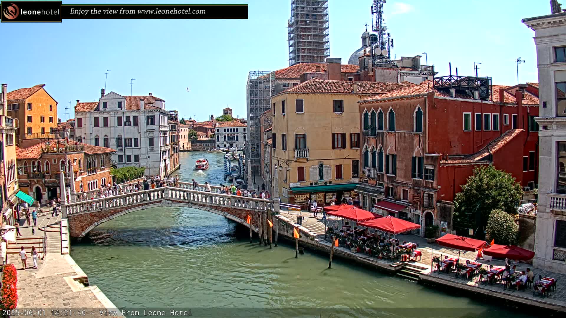 A sunny day in Venice shows a canal with a bridge, several buildings, and outdoor seating at a restaurant.