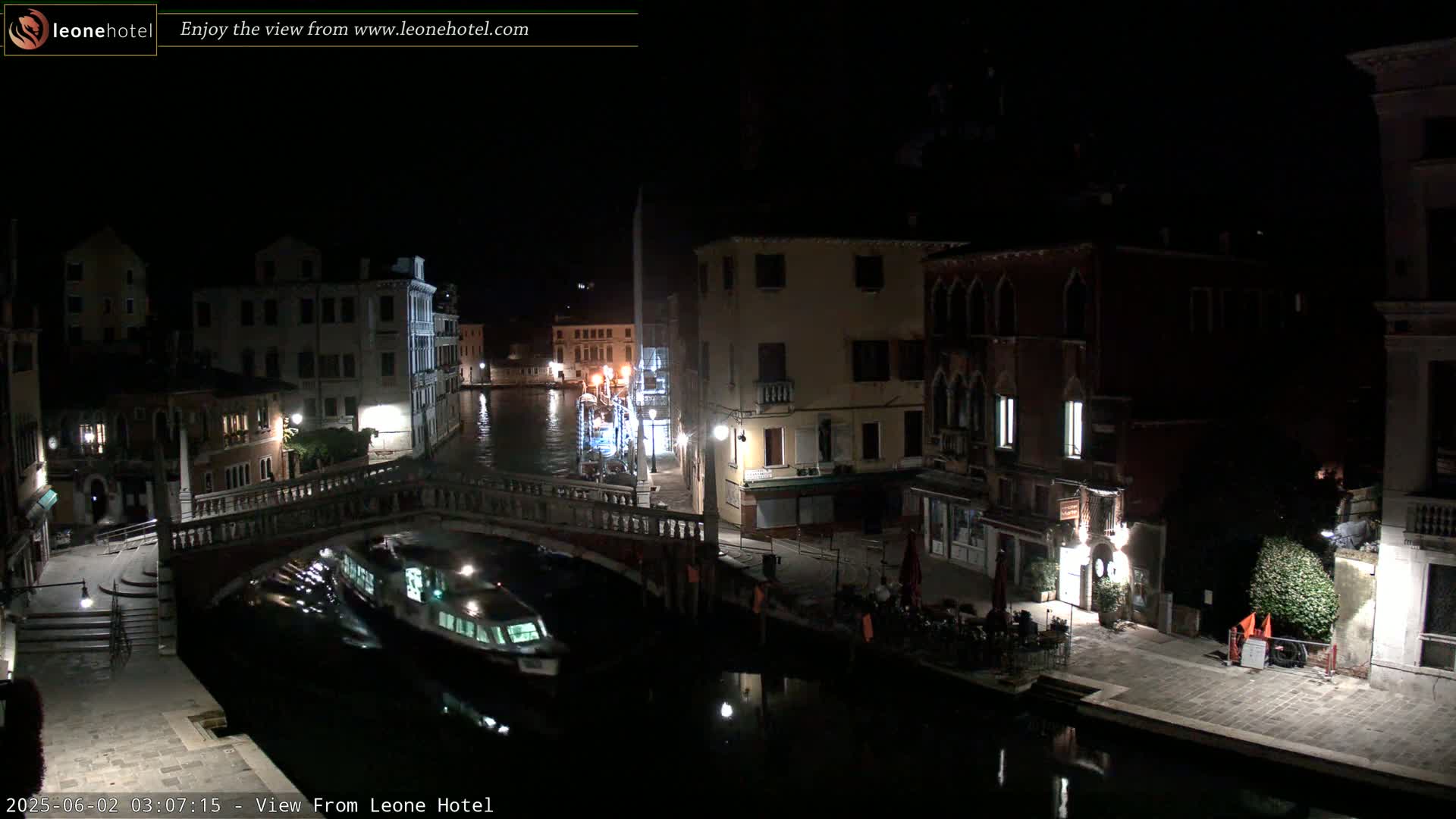 A nighttime view of a canal in Venice, Italy, shows a bridge, several buildings with lights on, and a boat on the water.
