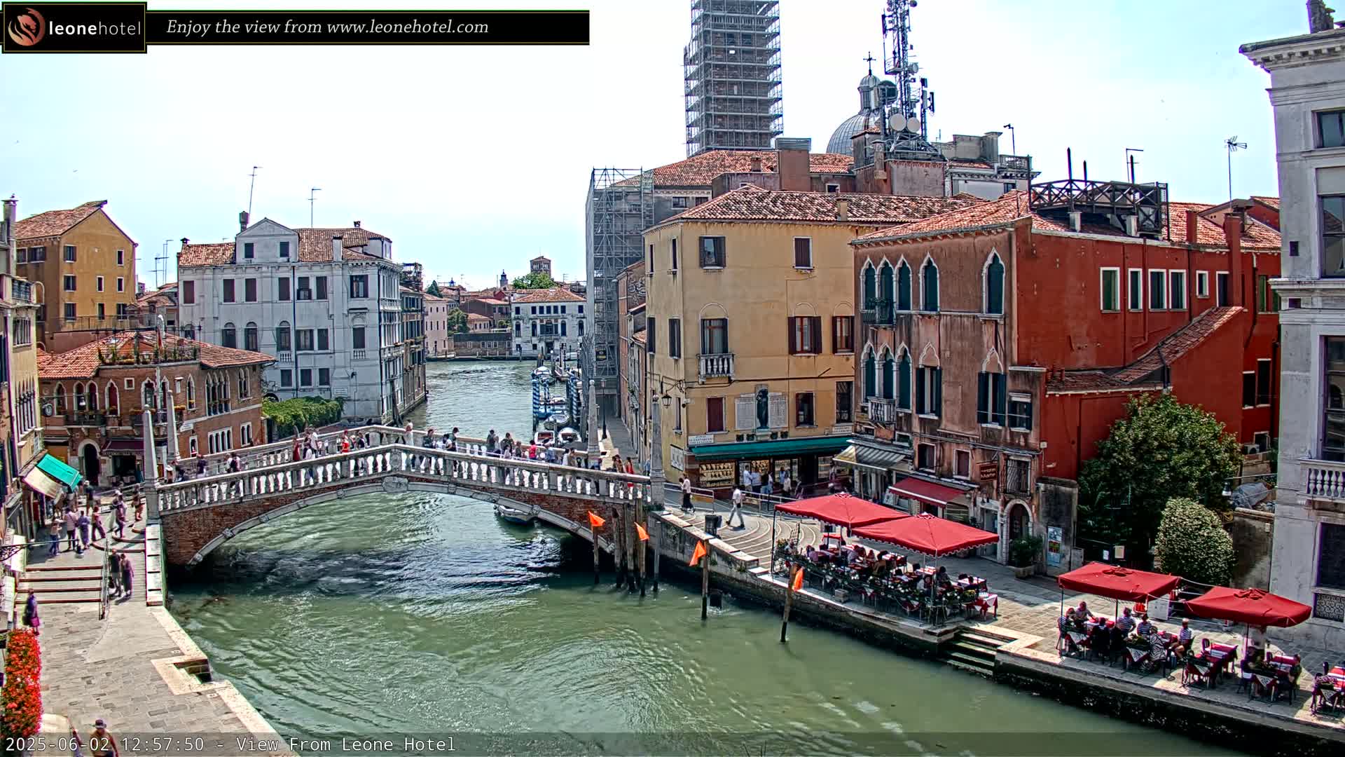 A sunny day in Venice shows a canal with a stone bridge, colorful buildings lining the waterway, and outdoor seating at restaurants along the canal.