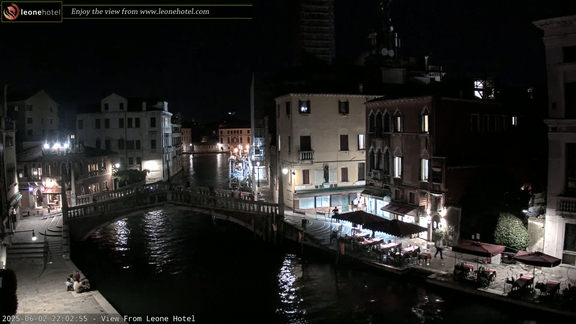 A nighttime view of a canal in Venice, showing bridges, buildings with illuminated windows, and outdoor seating at a restaurant.