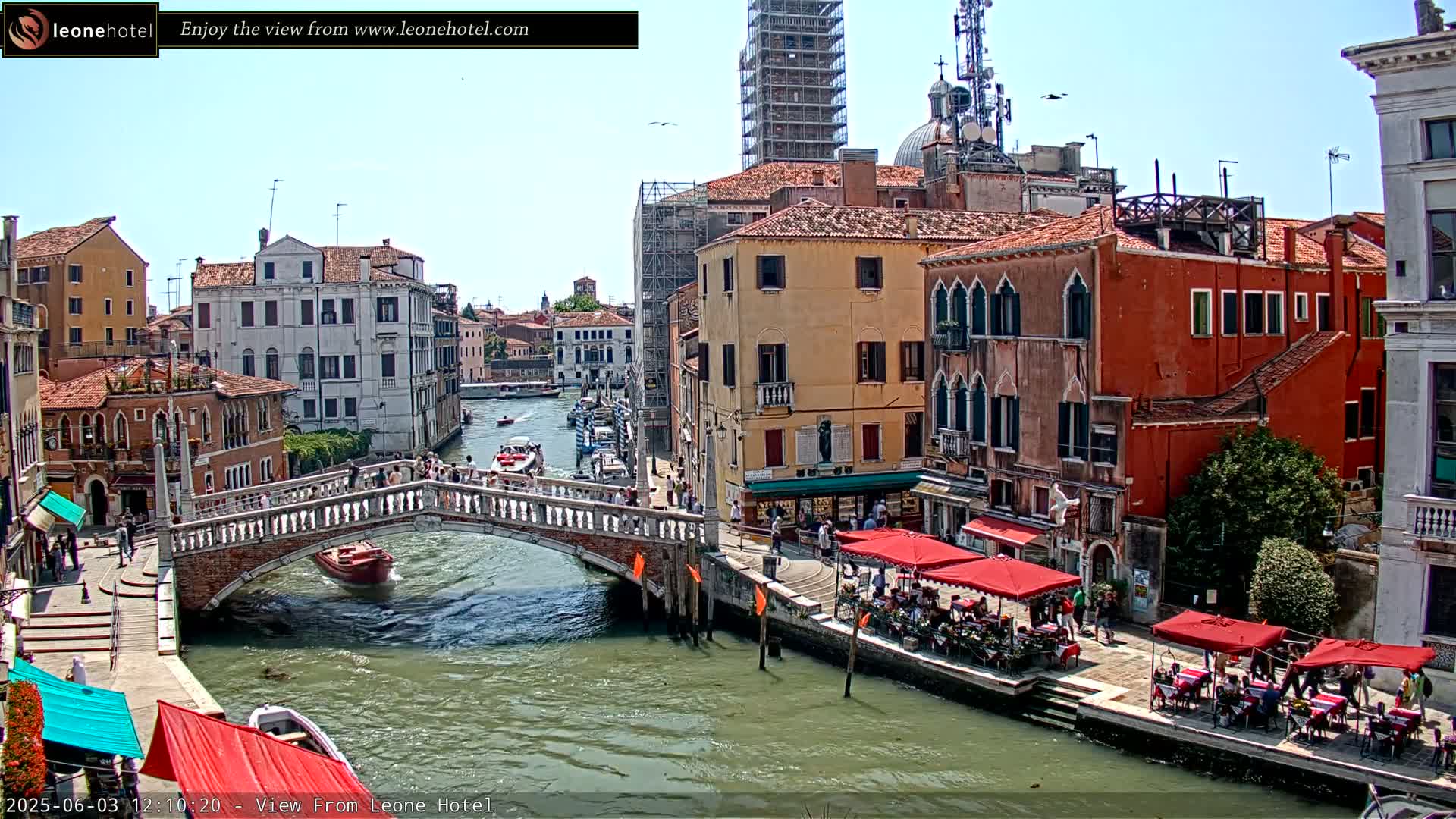 A sunny day in Venice shows a canal with boats, a stone bridge, and colorful buildings lining the waterway, with outdoor cafes set up along the canal.