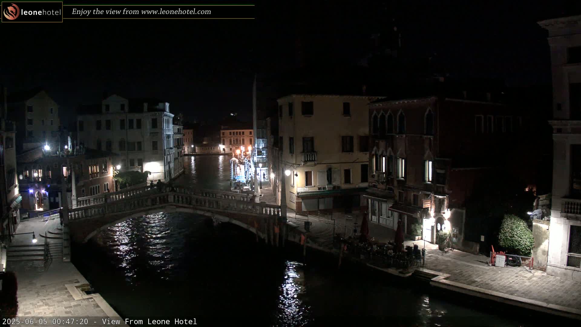 A nighttime view of a canal in Venice, Italy, showing a bridge, buildings lining the waterway, and some outdoor seating at a restaurant.