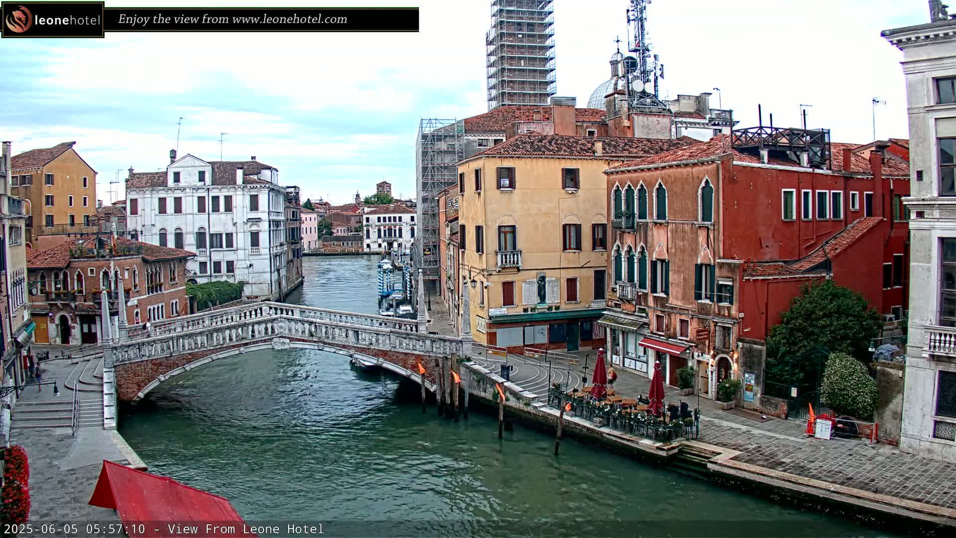 A stone bridge spans a canal in Venice, Italy, under a partly cloudy sky; colorful buildings line the waterway on either side.