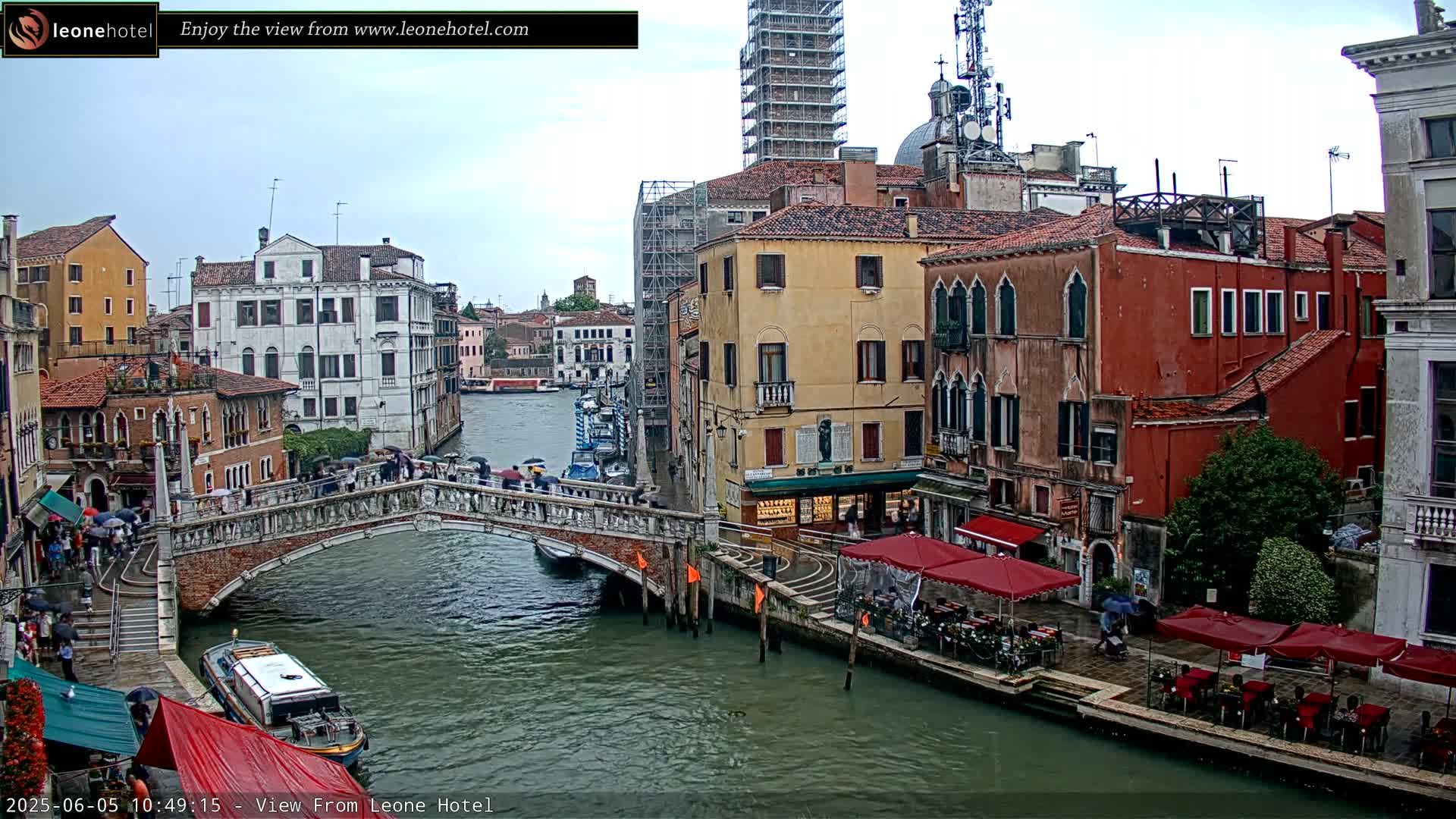 A view of a canal in Venice, Italy, on an overcast day, shows a bridge with pedestrians crossing, boats on the water, and buildings lining the canal.