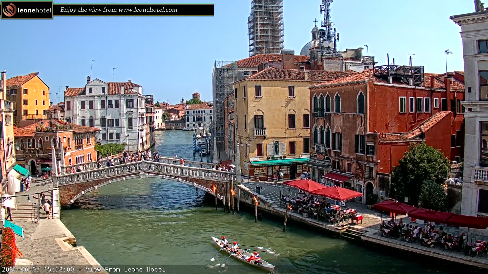 A sunny day in Venice shows a canal with a stone bridge, several colorful buildings, and a rowboat with people rowing.