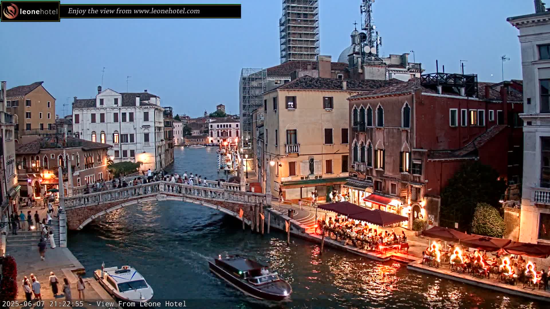 A twilight view of a Venetian canal, showing bridges, buildings, boats, and outdoor dining areas with people illuminated by warm lights.
