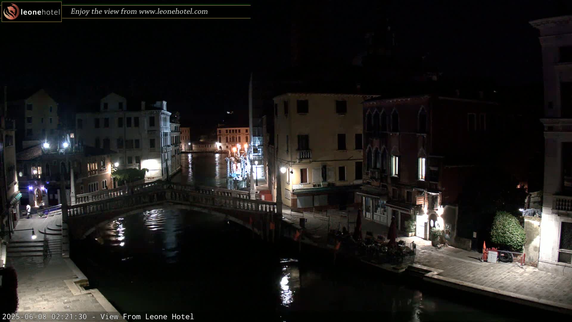 A nighttime view of a canal in Venice, Italy, shows several buildings along the waterway, a bridge, and a few people sitting outdoors.