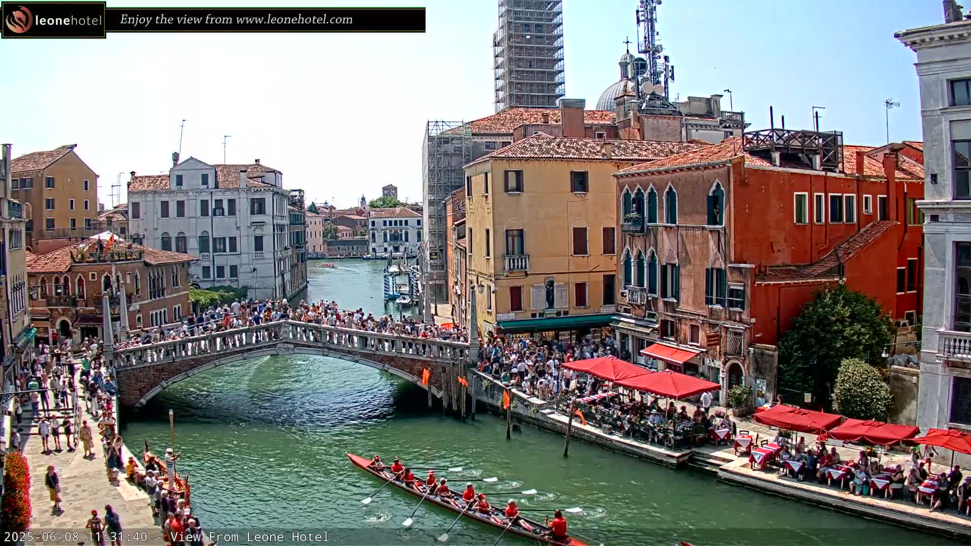 A crowded Venetian bridge overlooks a canal with a rowing team in a gondola and outdoor cafes lining the waterway under a sunny sky.