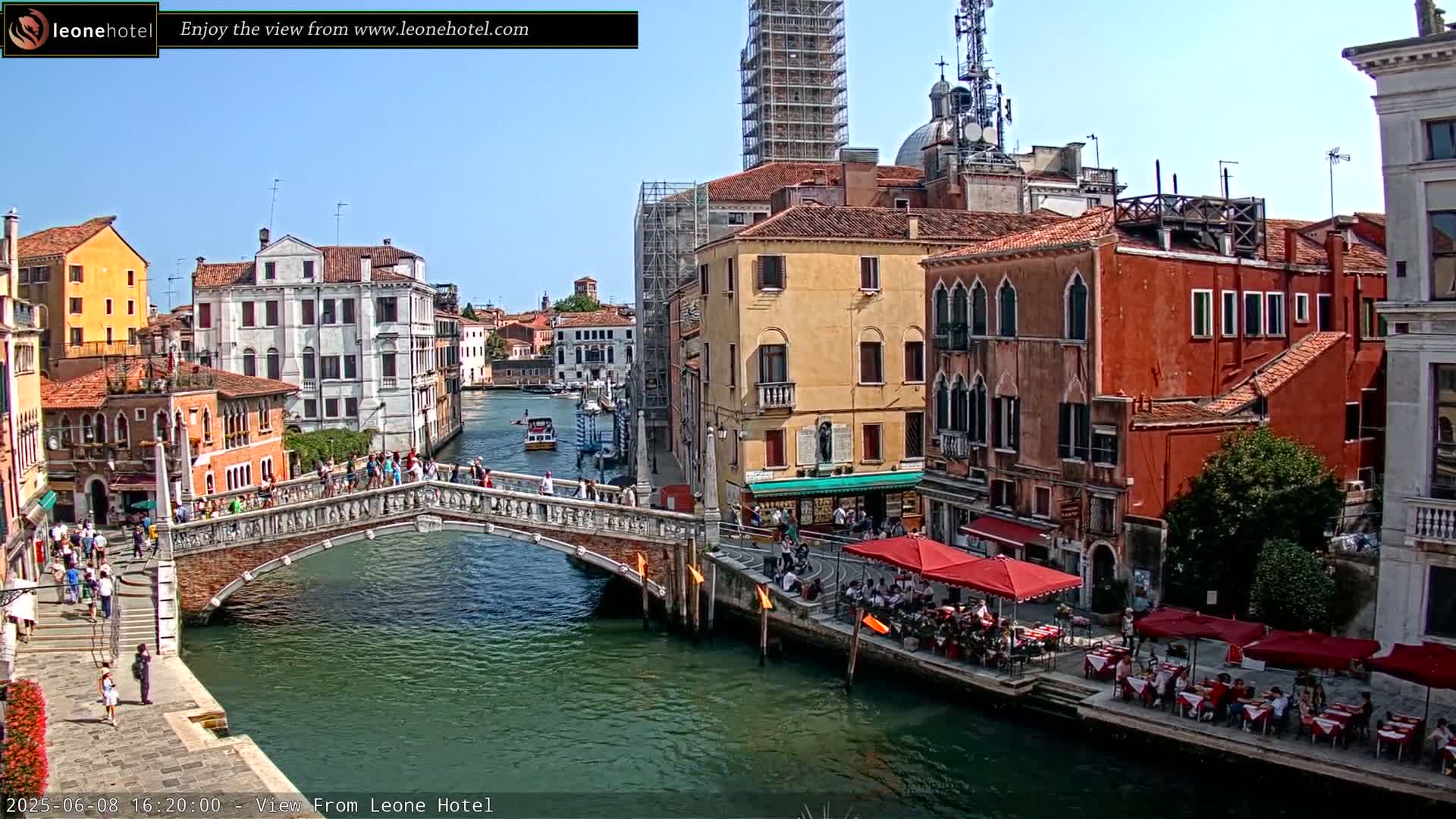 A sunny day in Venice shows a canal with a stone bridge, colorful buildings lining the waterways, and outdoor seating at waterfront restaurants.