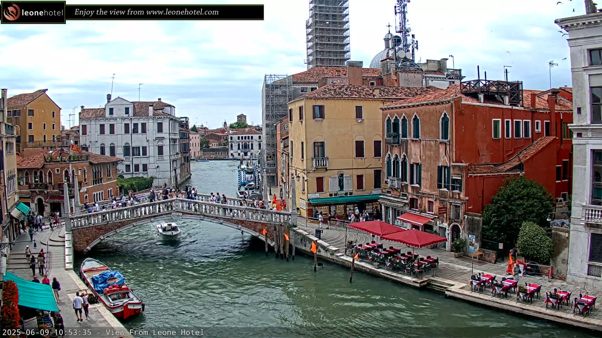 A canal in Venice, Italy, under a partly cloudy sky, shows a bridge with pedestrians, boats on the water, and buildings with outdoor seating along the canal.
