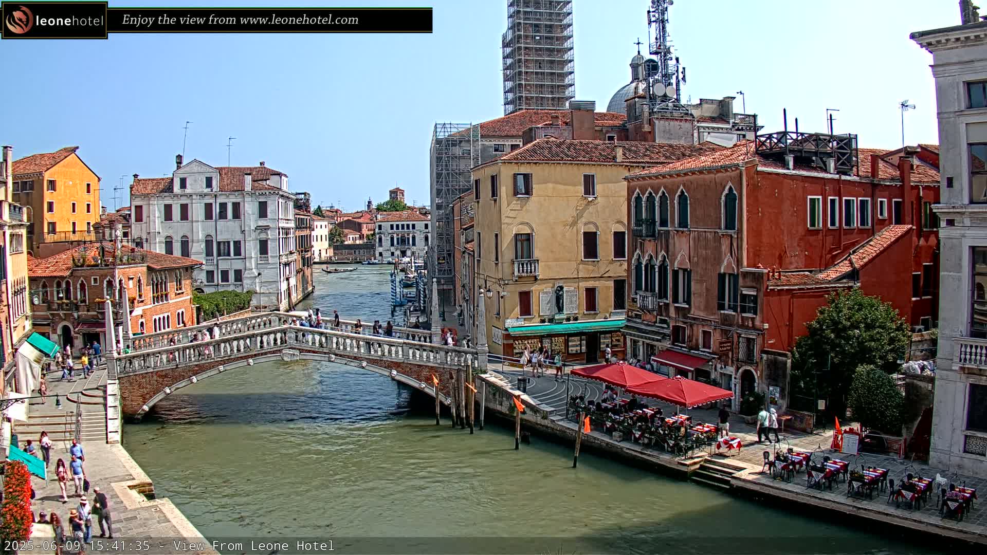 A sunny day in Venice shows a canal with a stone bridge, colorful buildings lining the waterway, and people walking on the bridge and along the canalside.