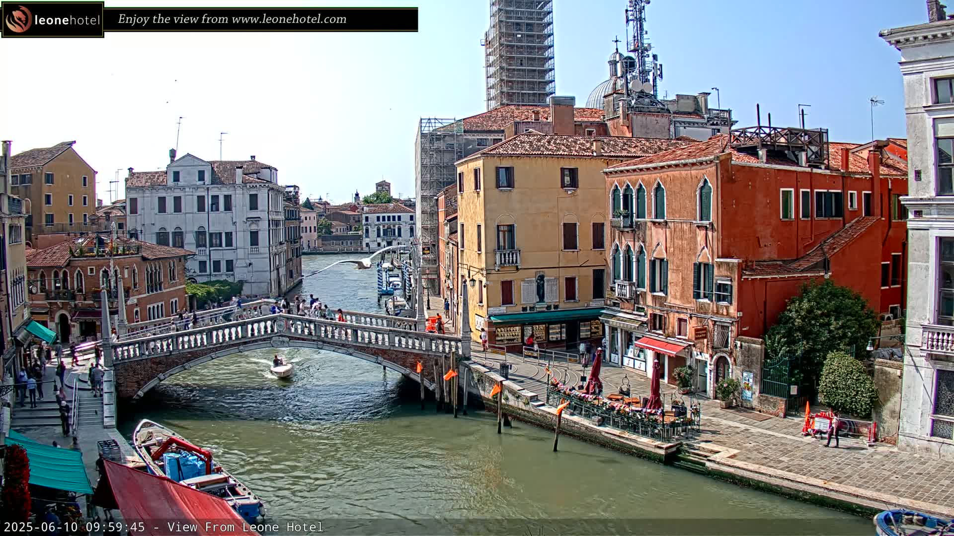 A sunny day in Venice shows a canal with a stone bridge, boats, and colorful buildings lining the waterway.