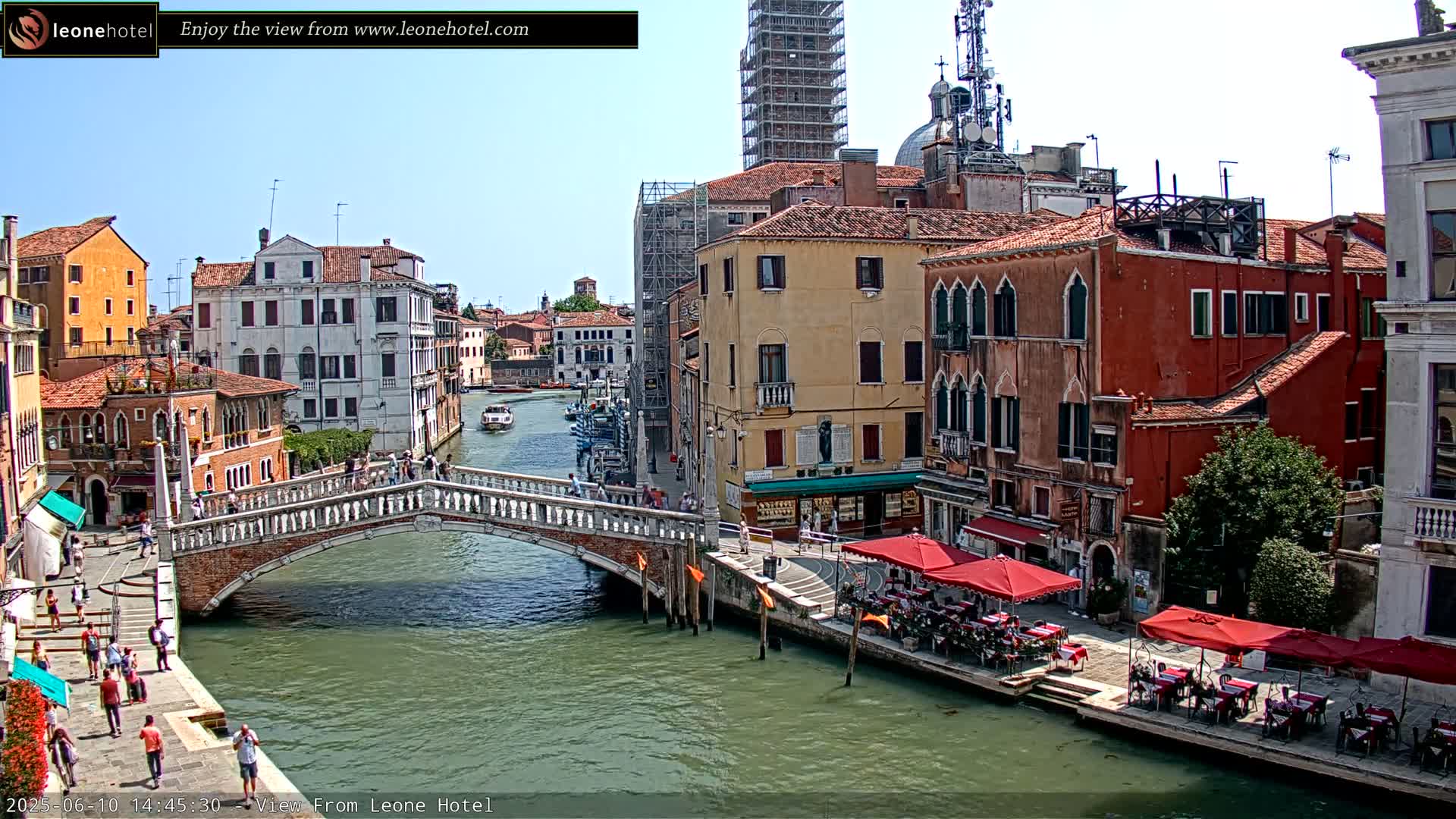 A sunny day in Venice shows a canal with several boats, a stone bridge, and colorful buildings with outdoor seating along the waterfront.