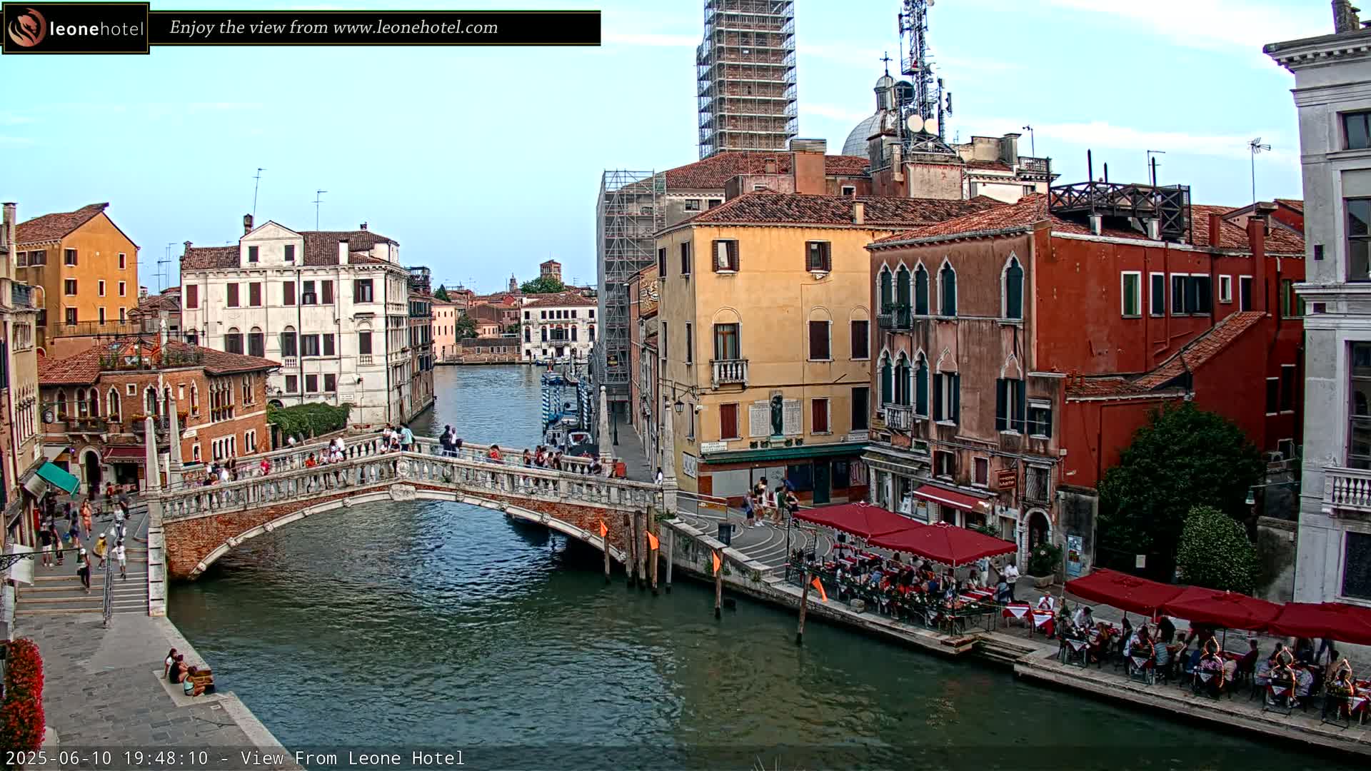 A bridge over a canal in Venice, Italy, under a mostly clear sky, is lined with people and outdoor restaurants.