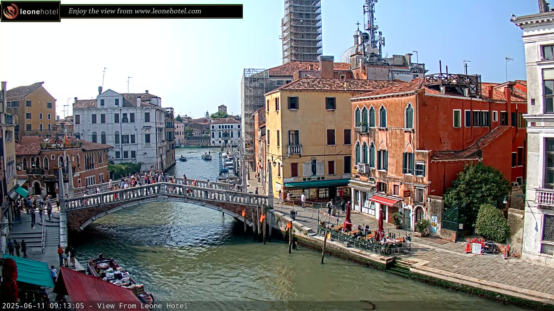 A sunny day in Venice shows a canal with boats, a stone bridge with people crossing it, and colorful buildings lining the waterway.