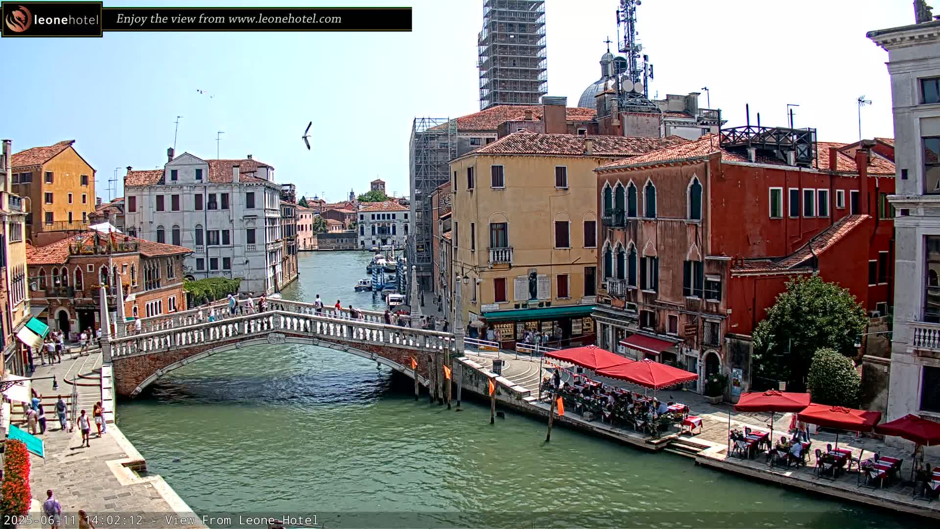 A sunny day in Venice shows a canal with a stone bridge, colorful buildings lining the waterway, and outdoor seating at cafés along the canal.