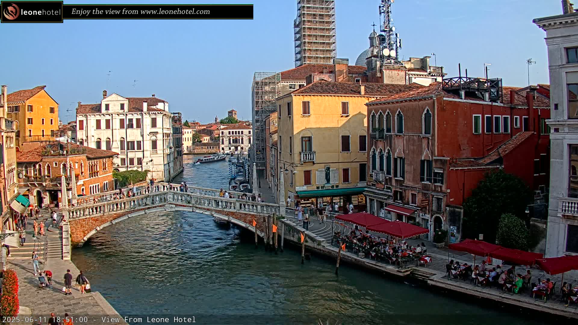 A canal in Venice, Italy, on a sunny day, is shown with colorful buildings lining the waterway, a stone bridge crossing the canal, and several outdoor cafes along the water's edge.