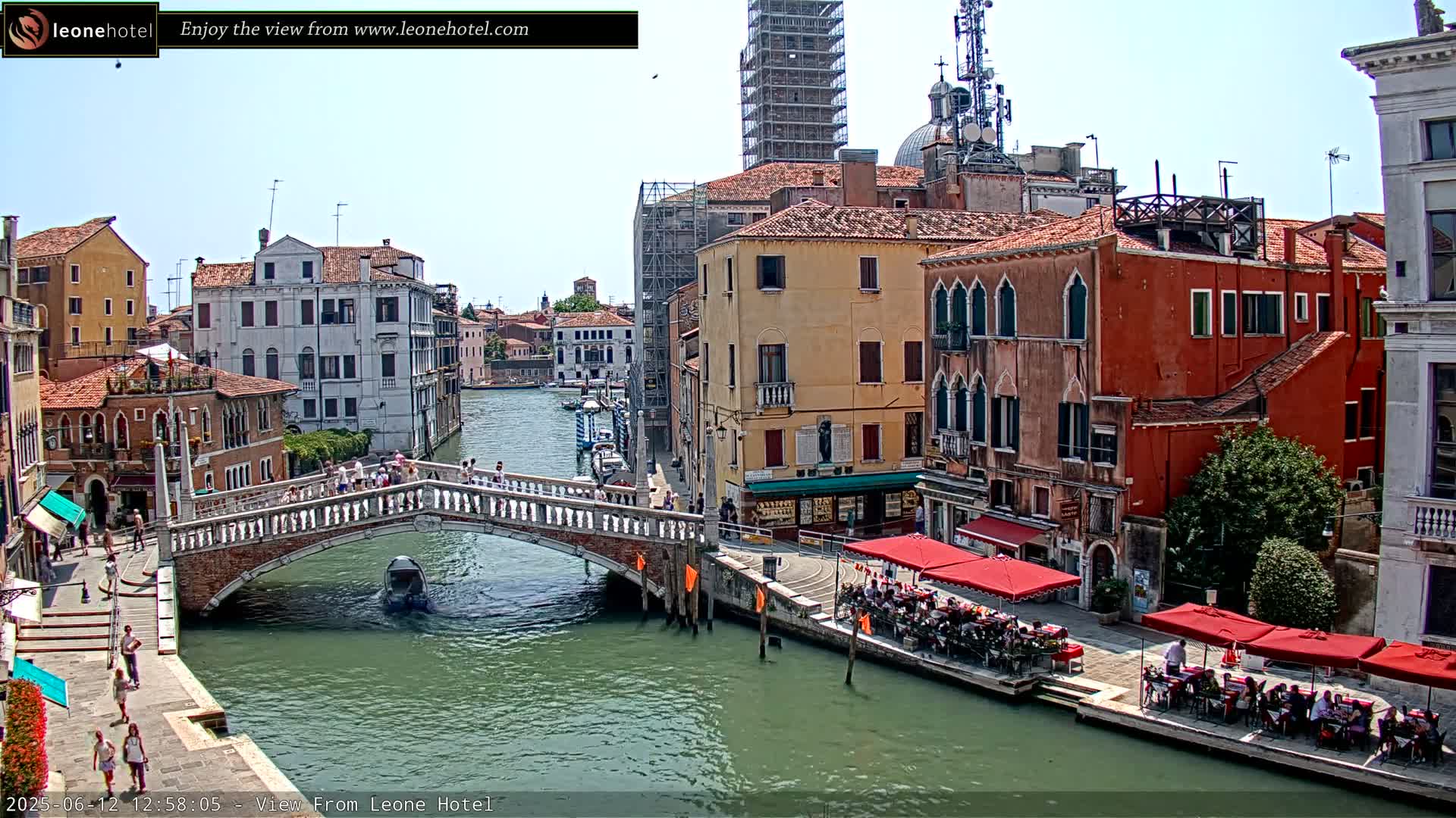 A sunny day in Venice shows a canal with colorful buildings, a bridge with pedestrians, and outdoor seating at a cafe.