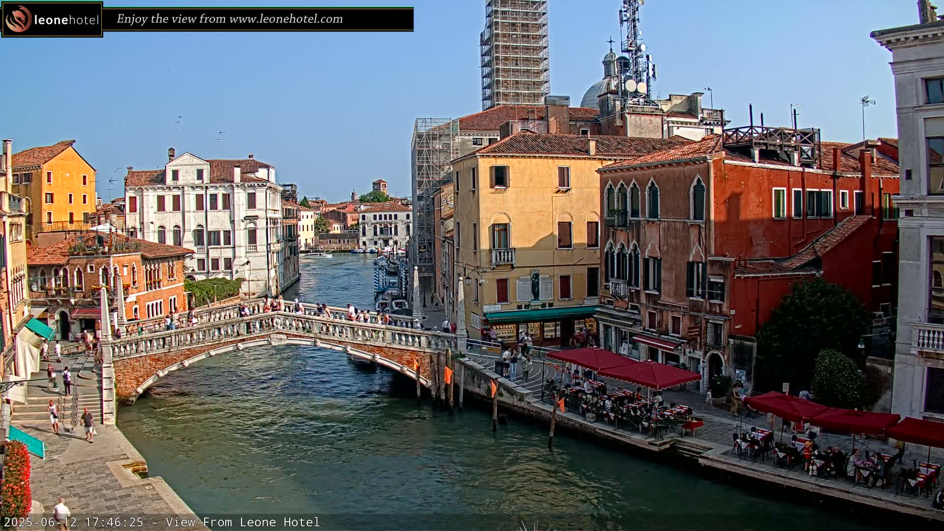 A sunny day in Venice shows a canal with a stone bridge, lined with colorful buildings, some with outdoor seating, and scaffolding on a tall building in the background.